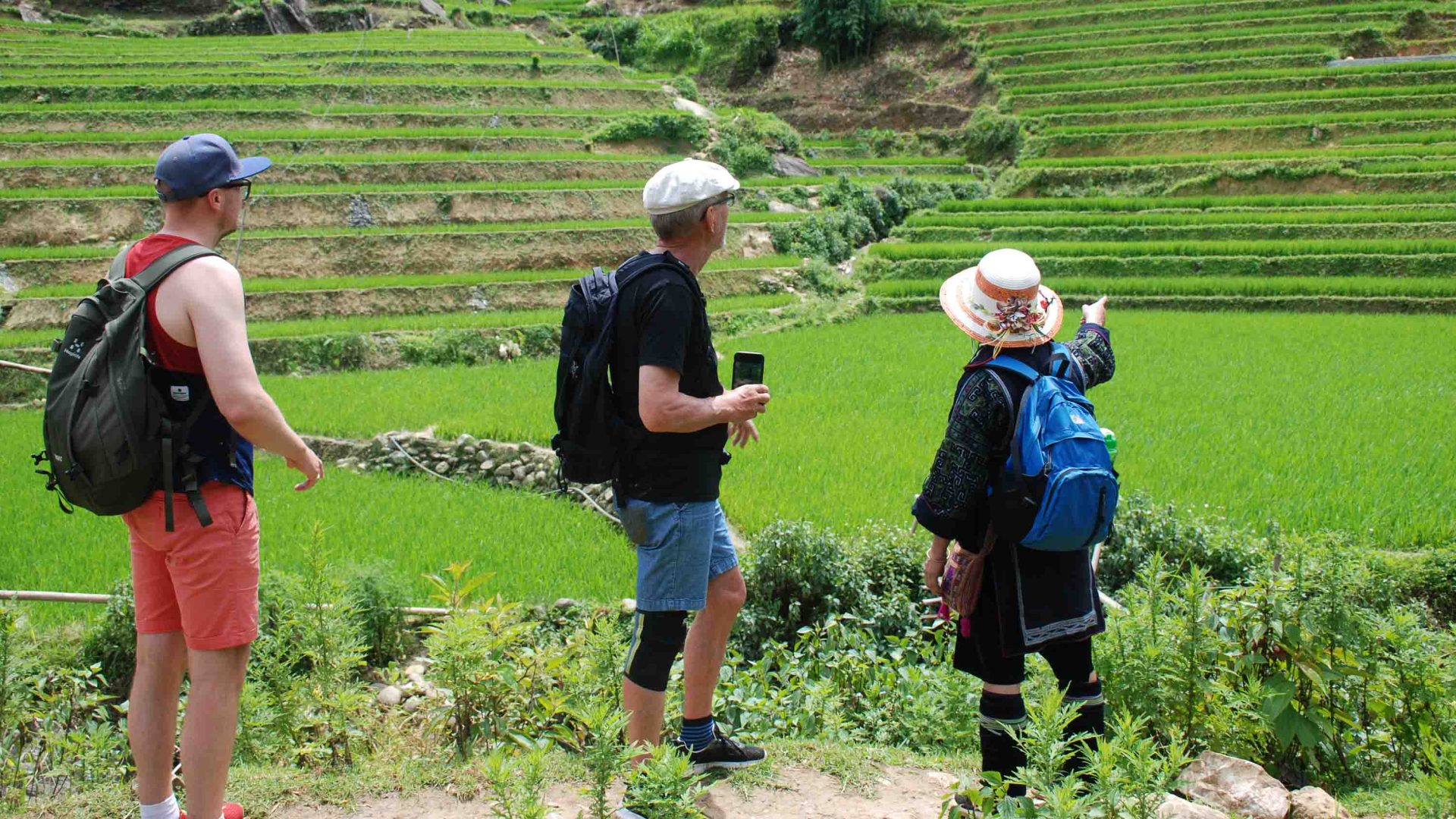 Two men listen to a local woman who points to something in green hills.