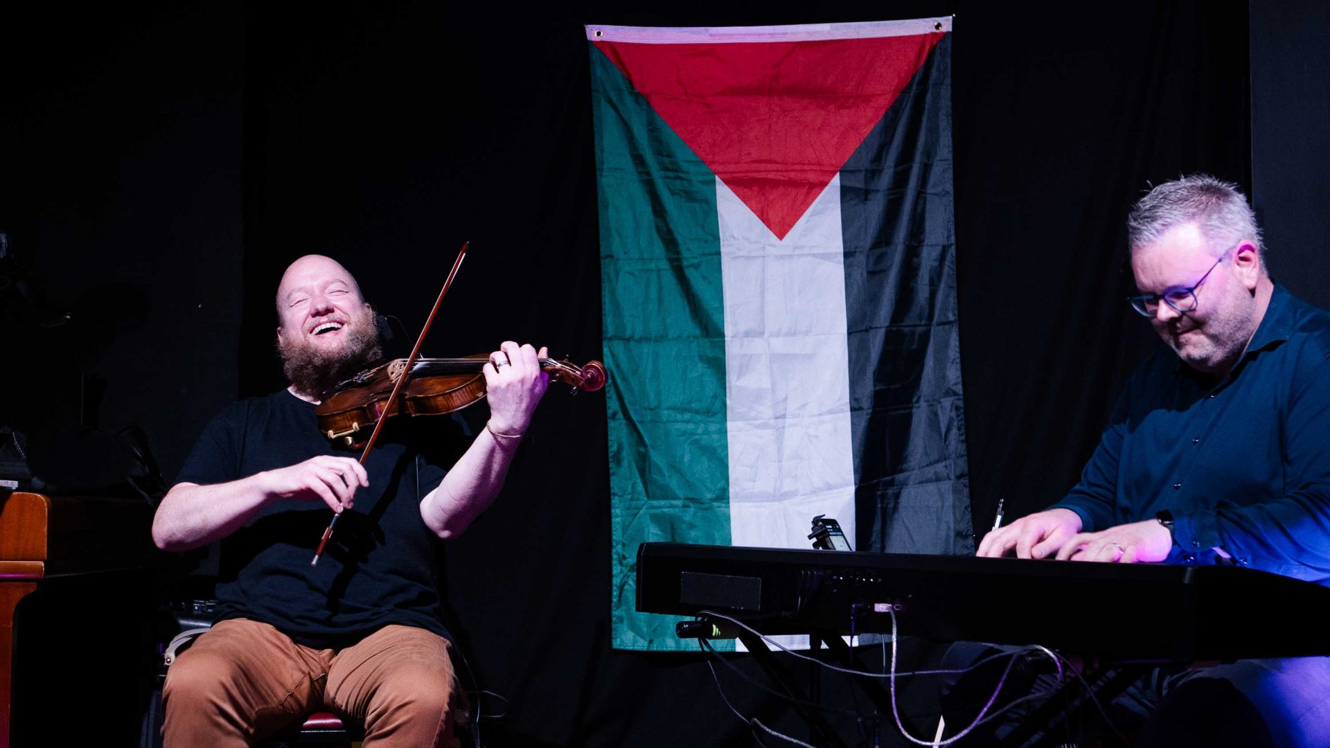 Two performers on stage with a Palestinian flag in the background.