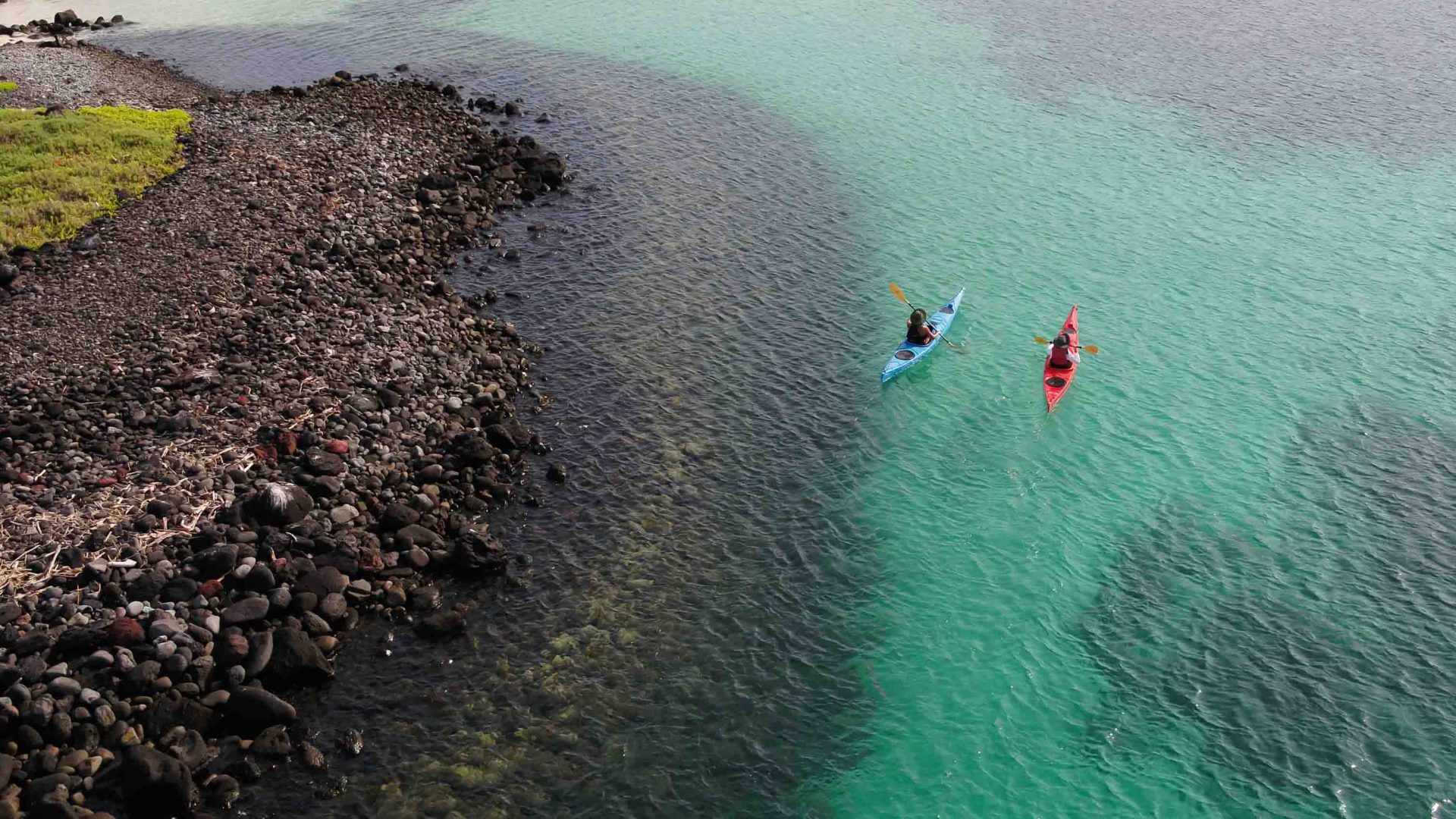 A drone photo of two people kayaking not far from shore in very clear blue water.