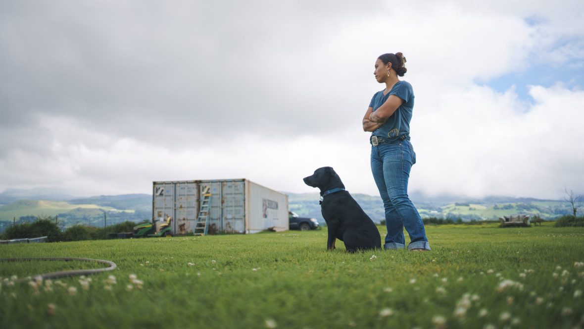 Woman wearing jeans stands beside black dog, they both look the same direction.