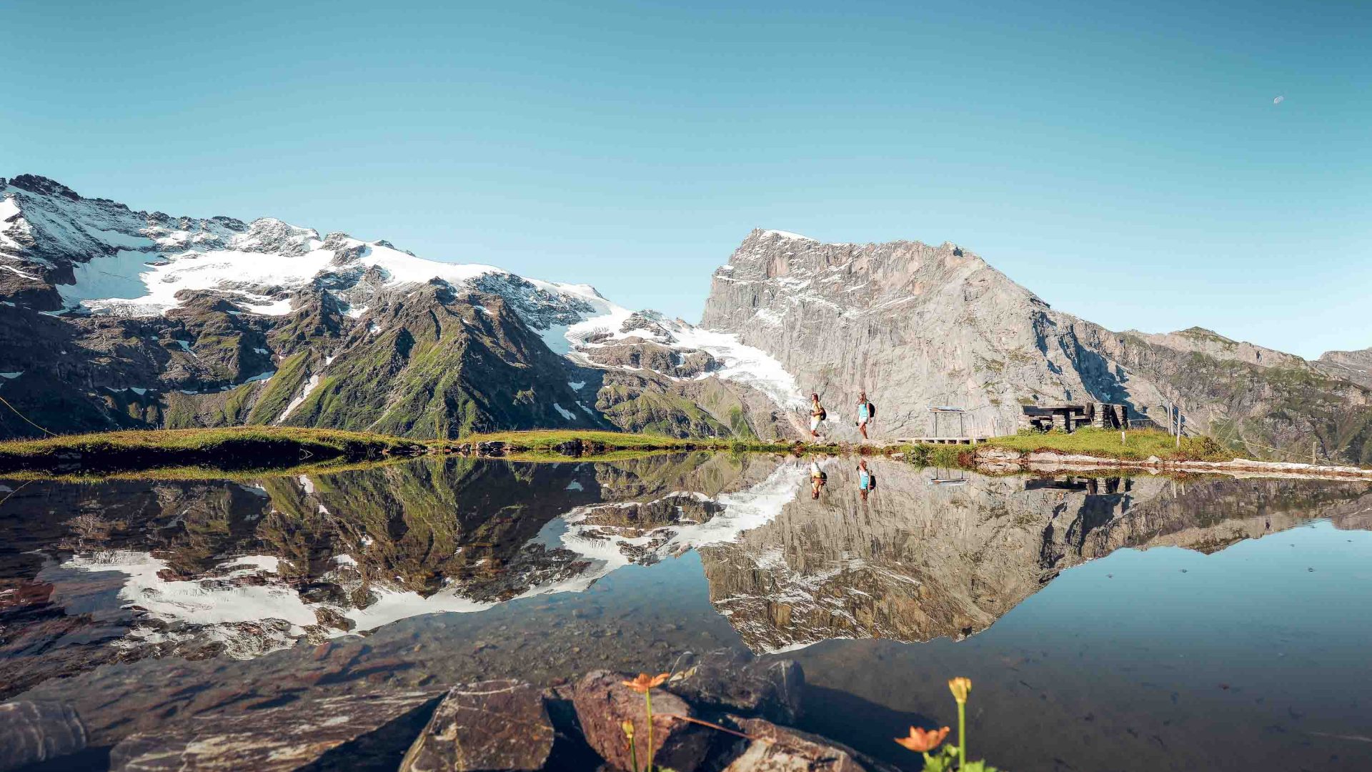 Mountains are reflected in a lake that has two people walking on its shore.
