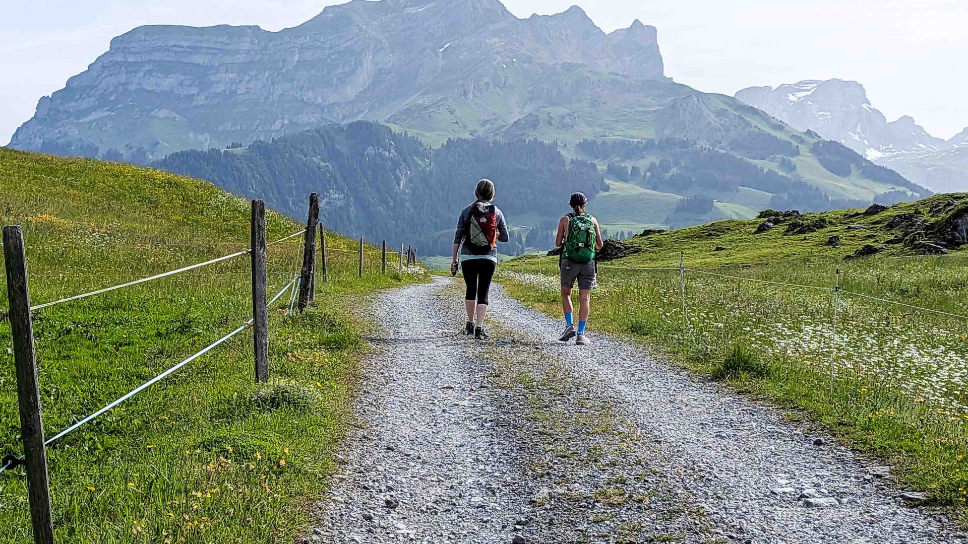 Two people walk along a path that leads to mountains.