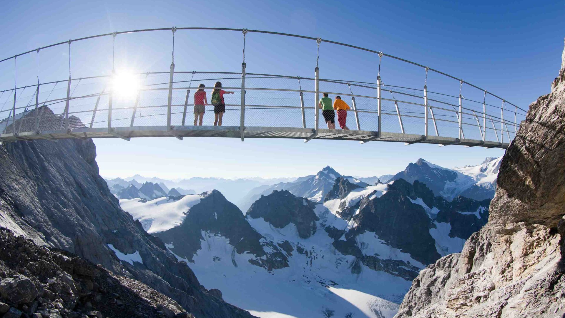 People stand on a bridge overlooking snowy mountains.