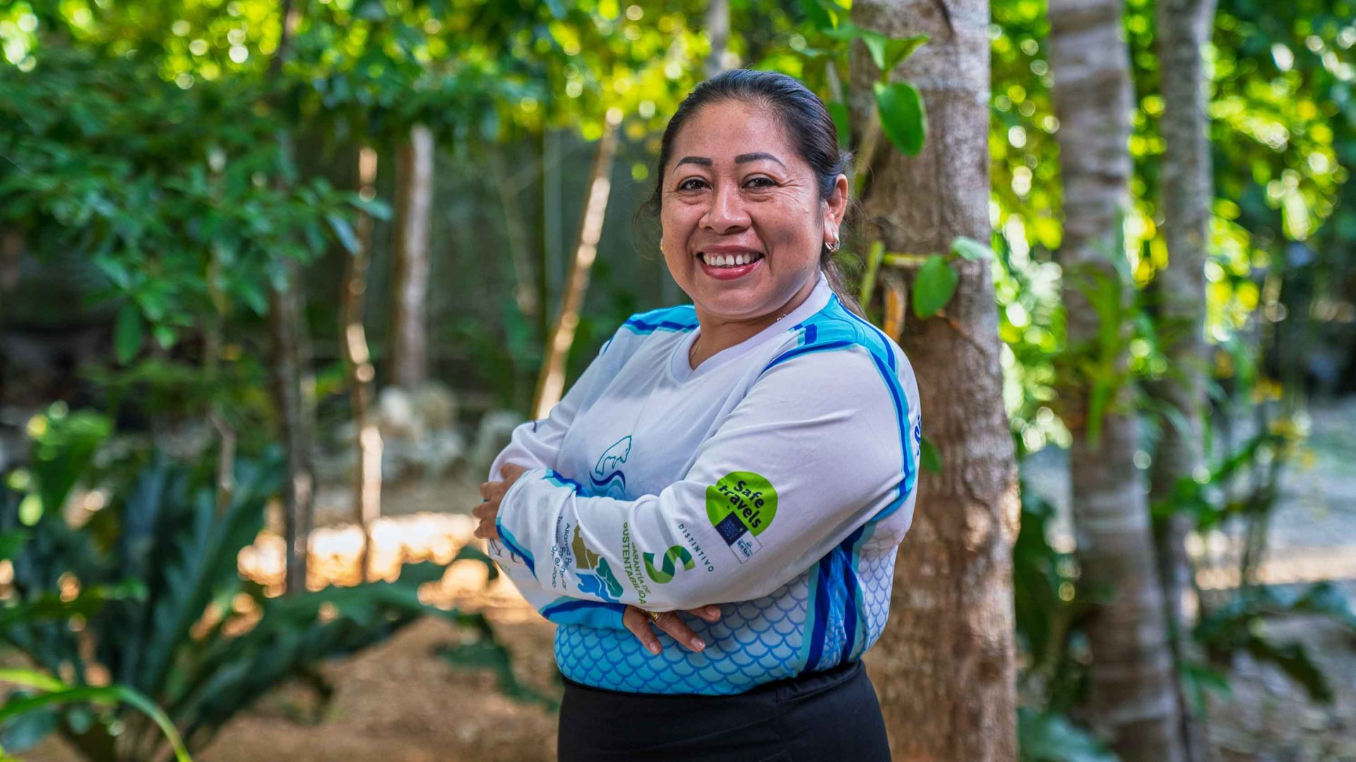 A woman stands with her arms crossed against her chest and a big smile. She is in a forest setting.
