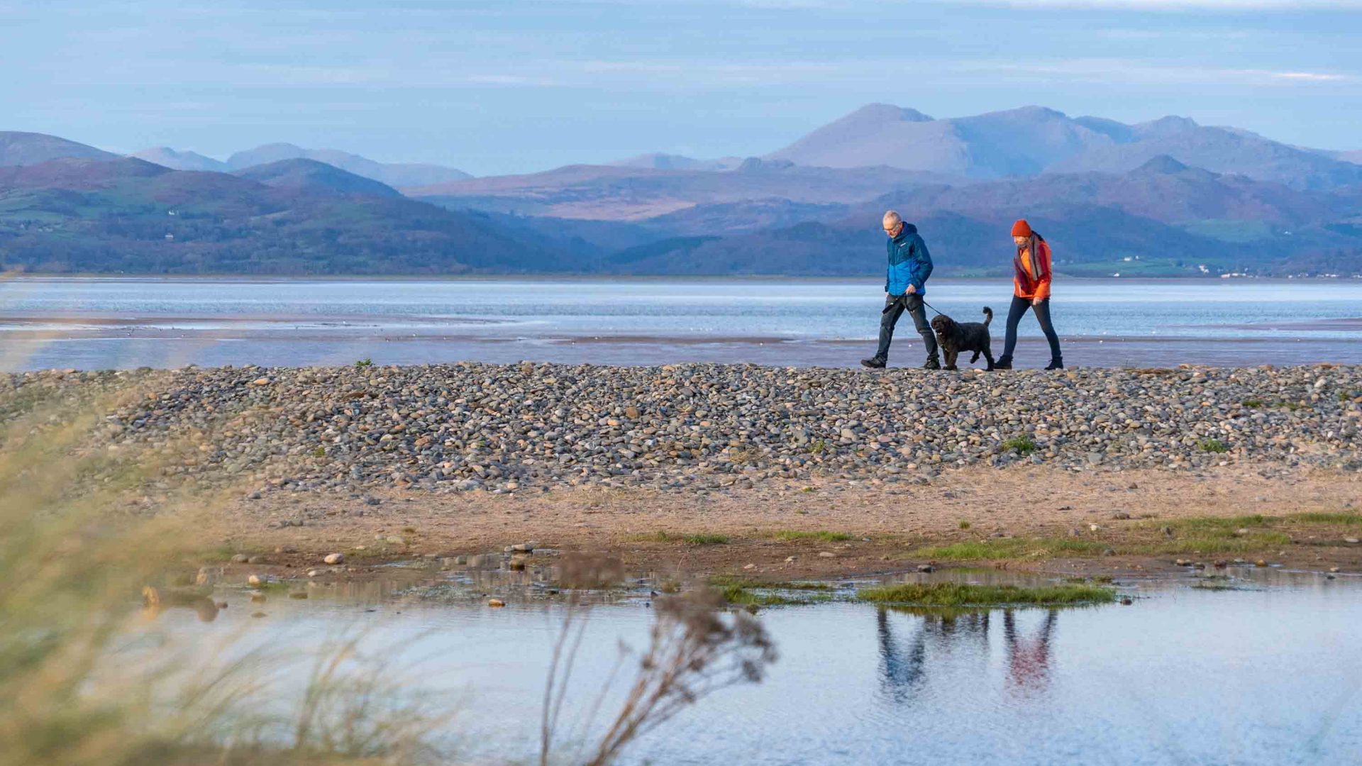 Two people walk along a lake with mountains in the background.