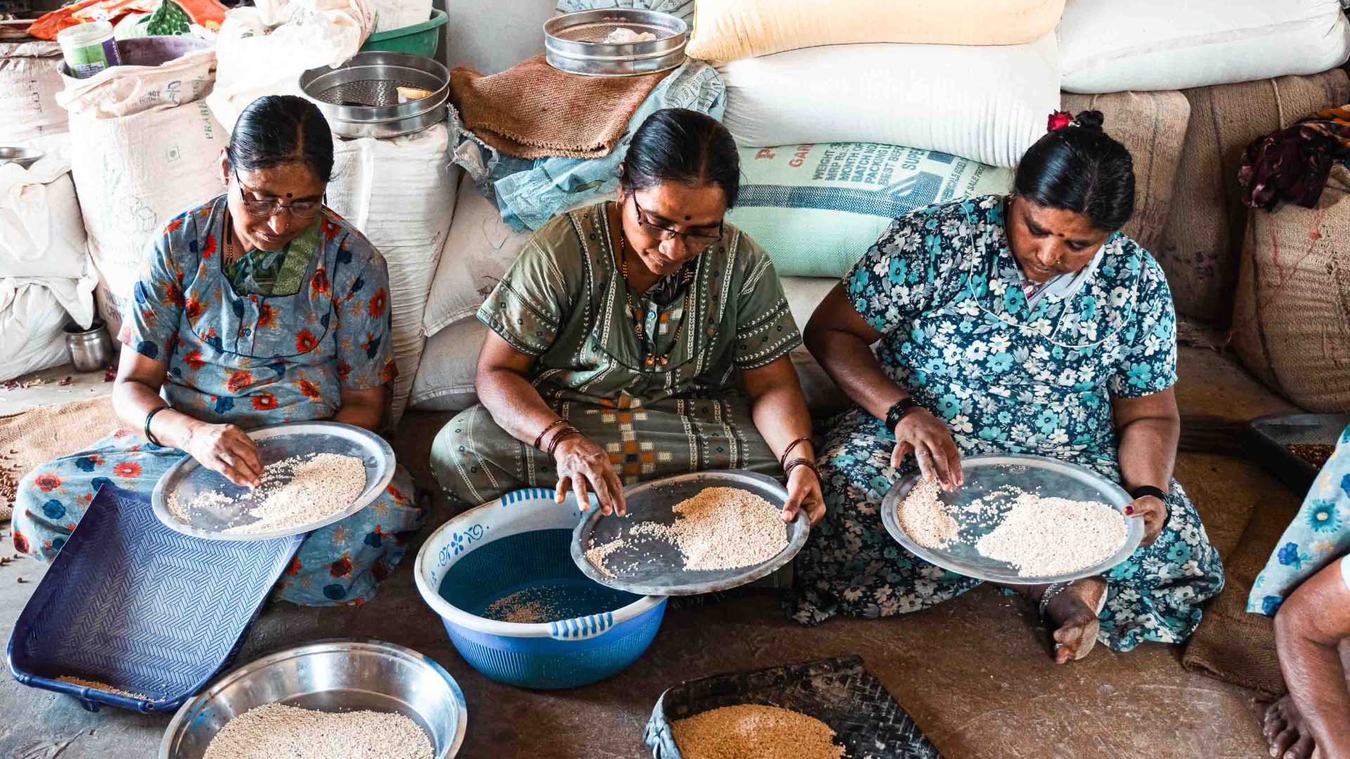 Three women sort through grains while seated on the ground.