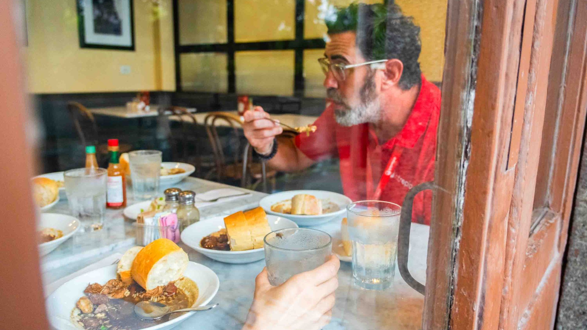 A man in a red Intrepid Travel t short is seen through a window bringing a fork to his mouth. There are plates of gumbo on the table in front of him.