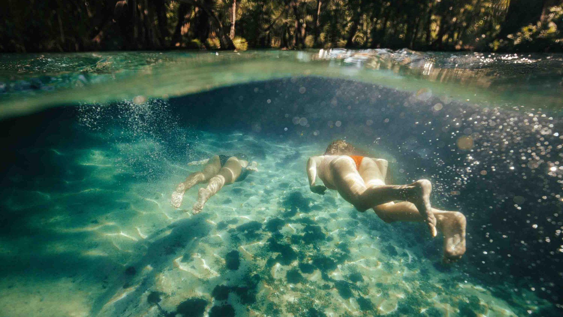 Two people swim through turquoise shallow waters with a sandy sea bed.