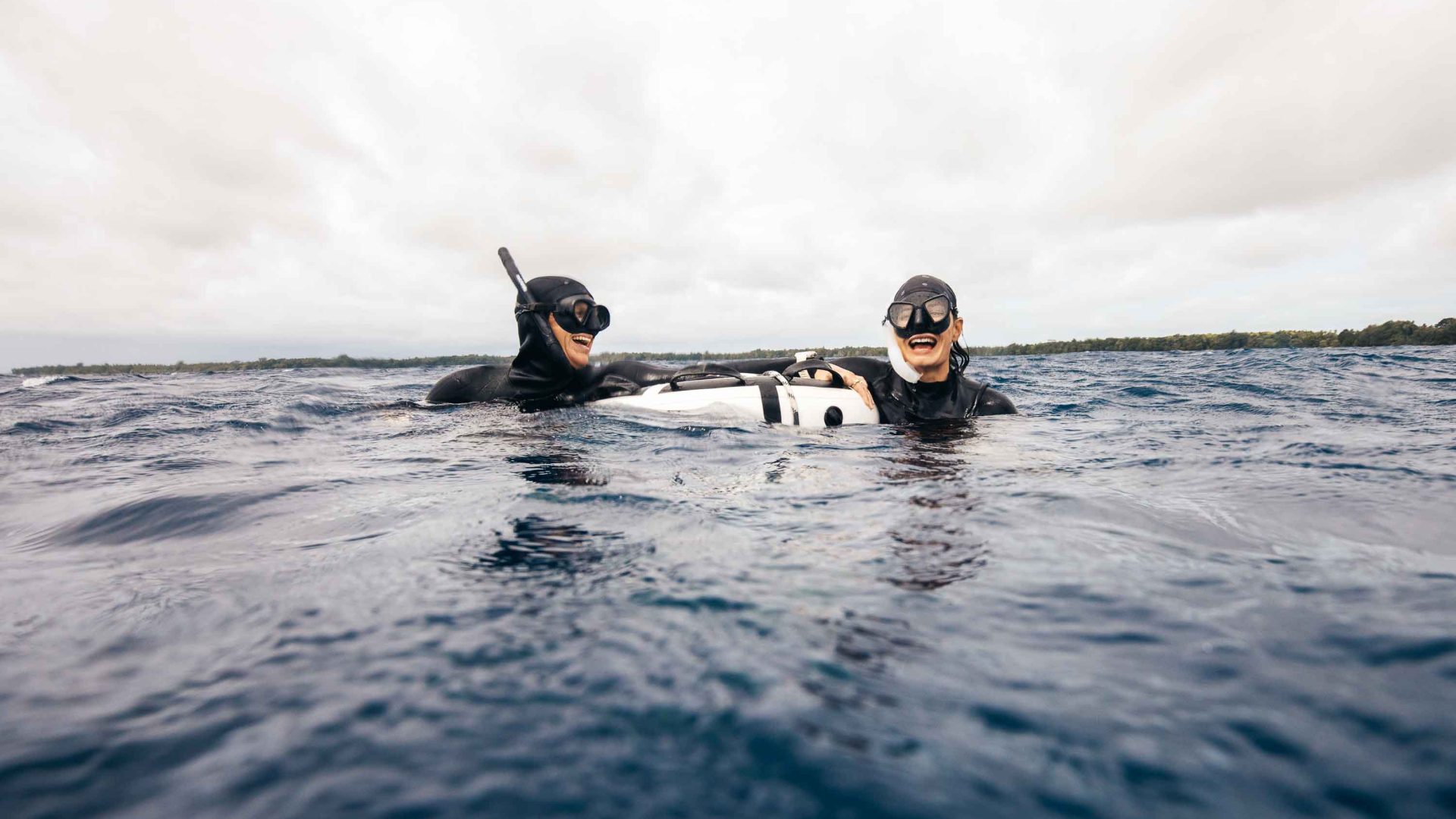 Two women laugh together while holding a buoy on the surface of the water. They are in diving wetsuits. with snorkels.