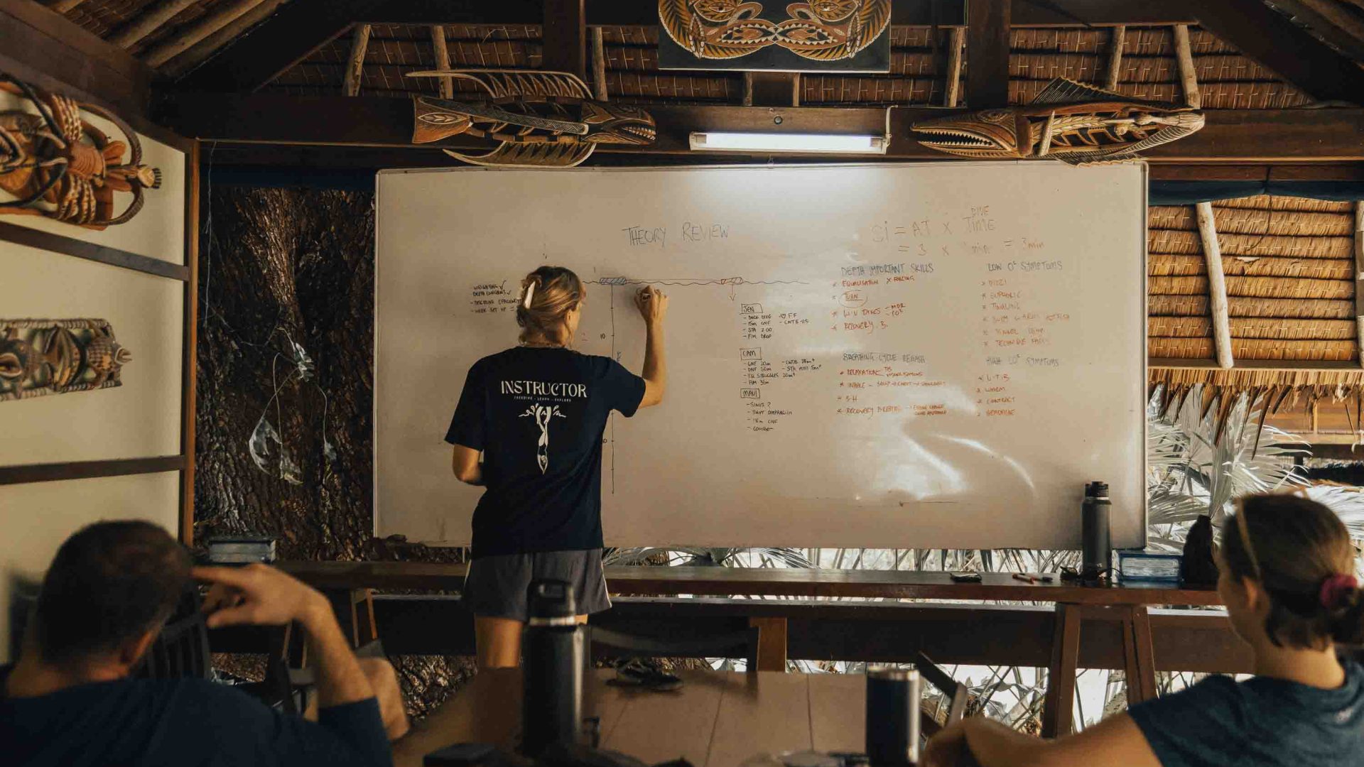 A woman writes on a white board while students listen. They are in a rustic building with bamboo walls.