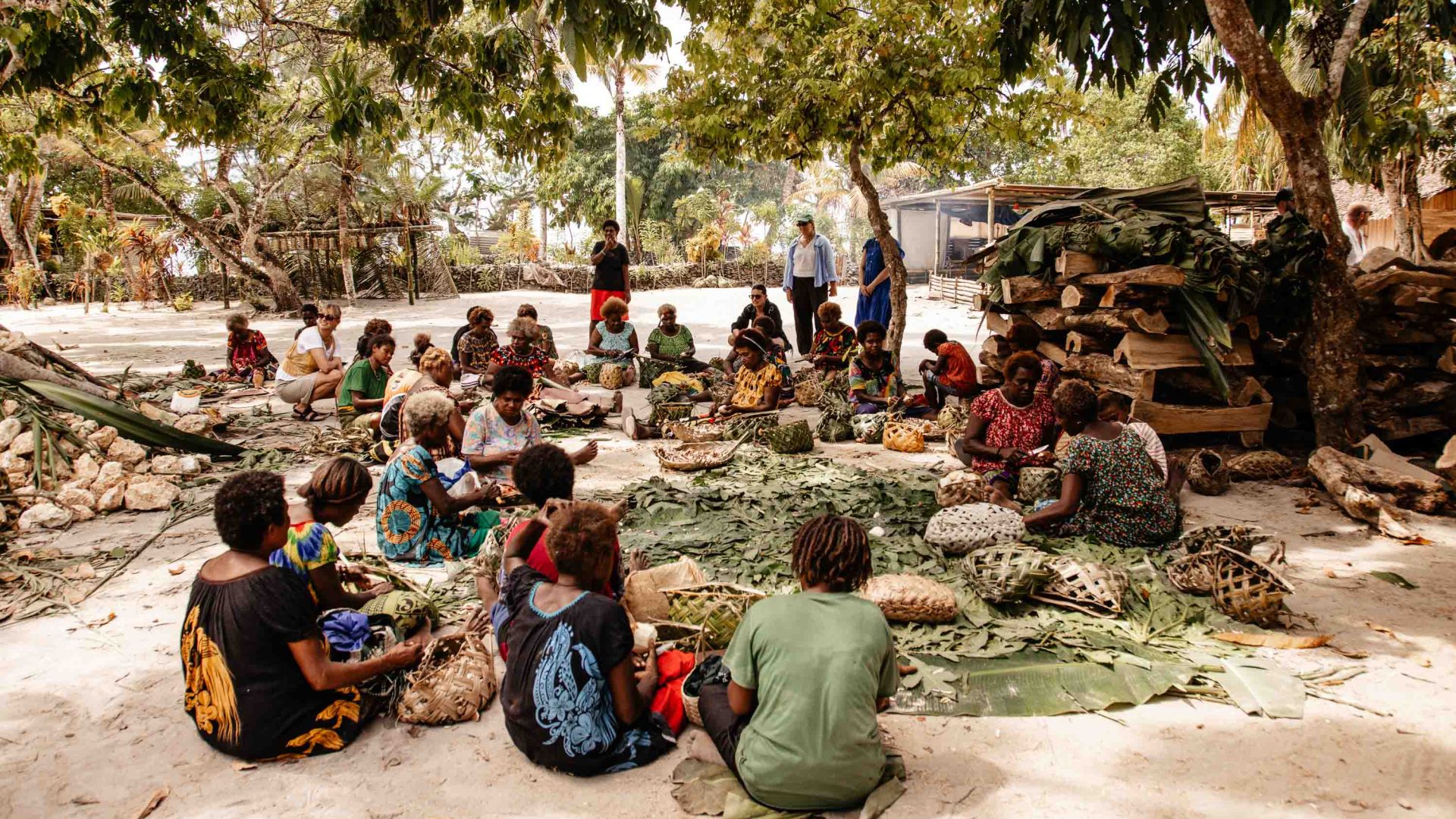 A group of local people sit on the ground outside, with green leaves for weaving in front of them.