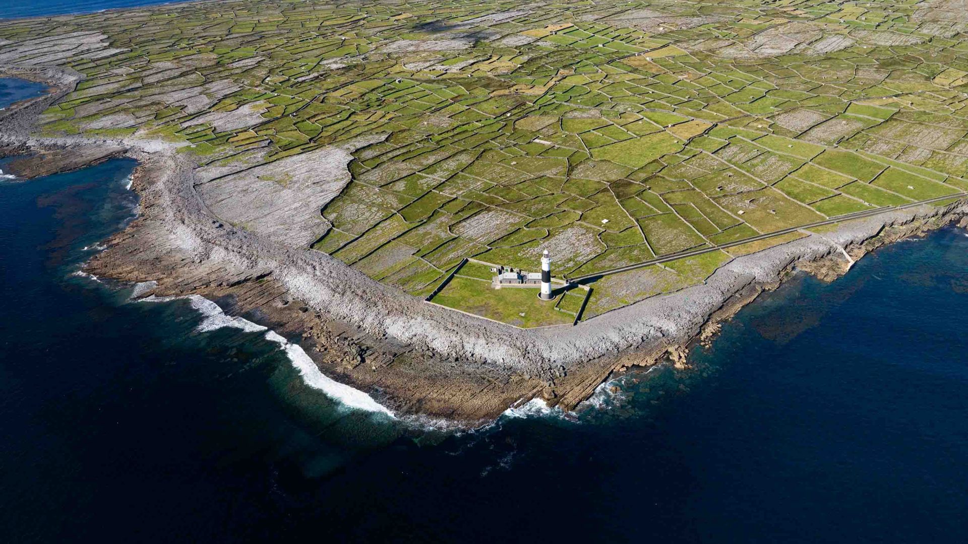 A drone photo of an island, showing a lighthouse at one end and a smattering of houses on the other end.