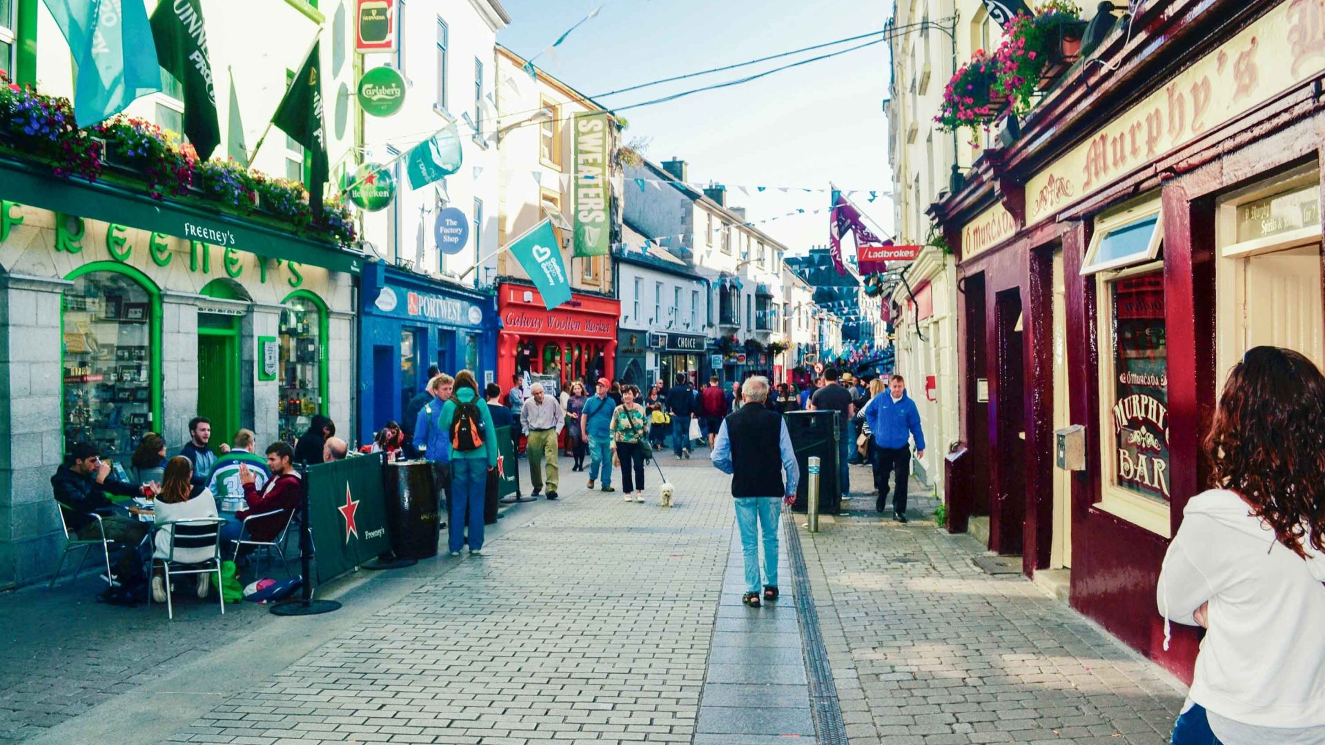 People walk through a pedestrian street lines with shops and Irish flags.