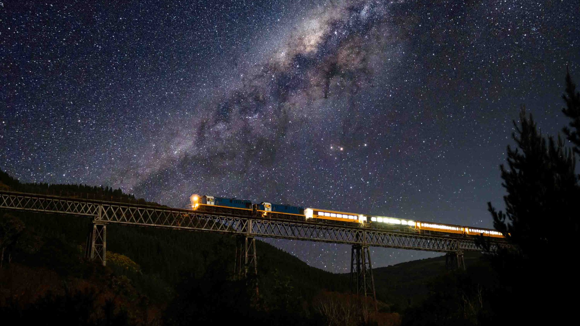 A train crosses an iron bridge at night under a clear, starry sky with a visible Milky Way galaxy. The bridge is lit with yellow lights, and the scene captures the contrast between the starry sky and the structural bridge.