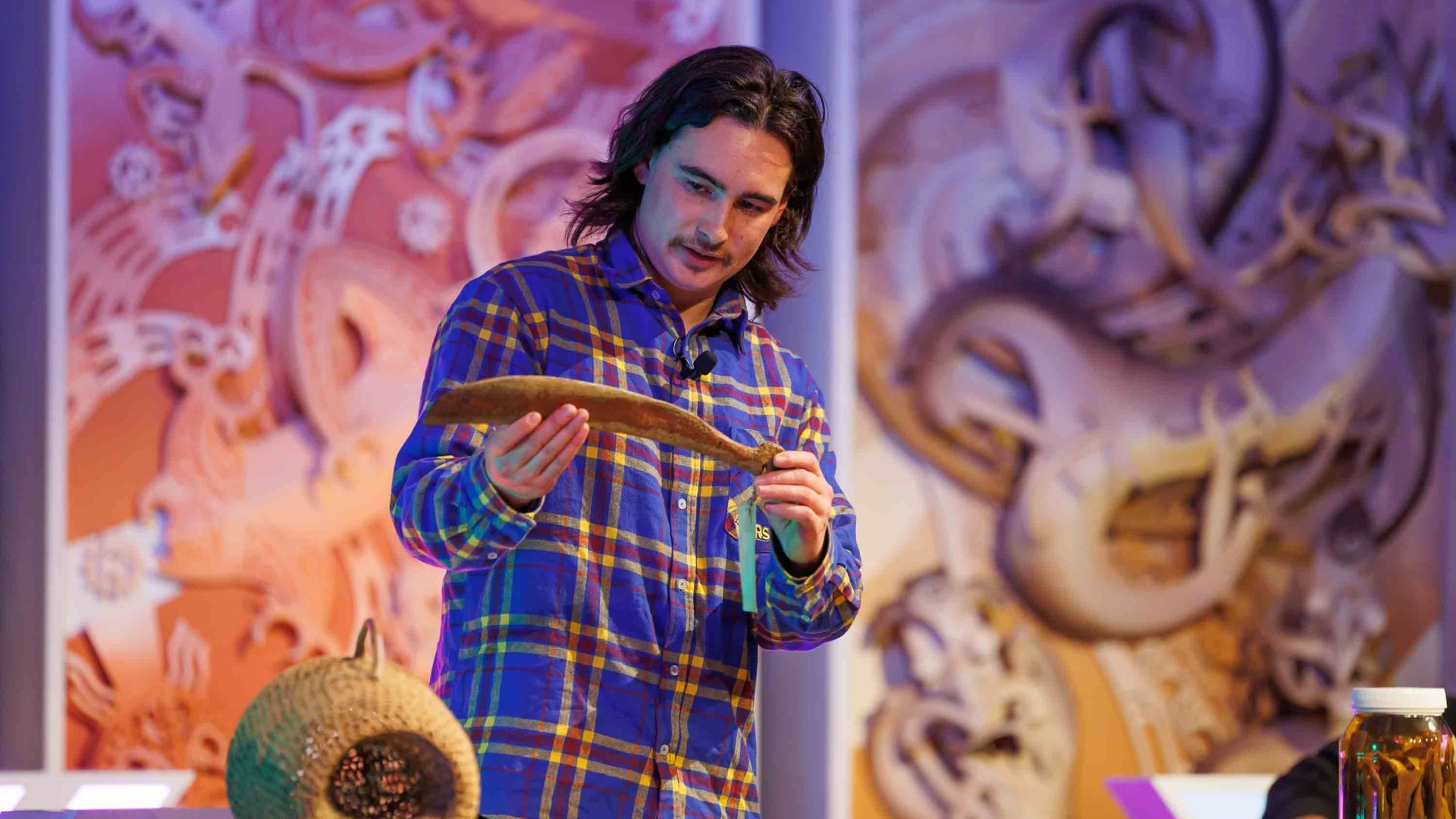 A man on a stage holds something wooden as he speaks.