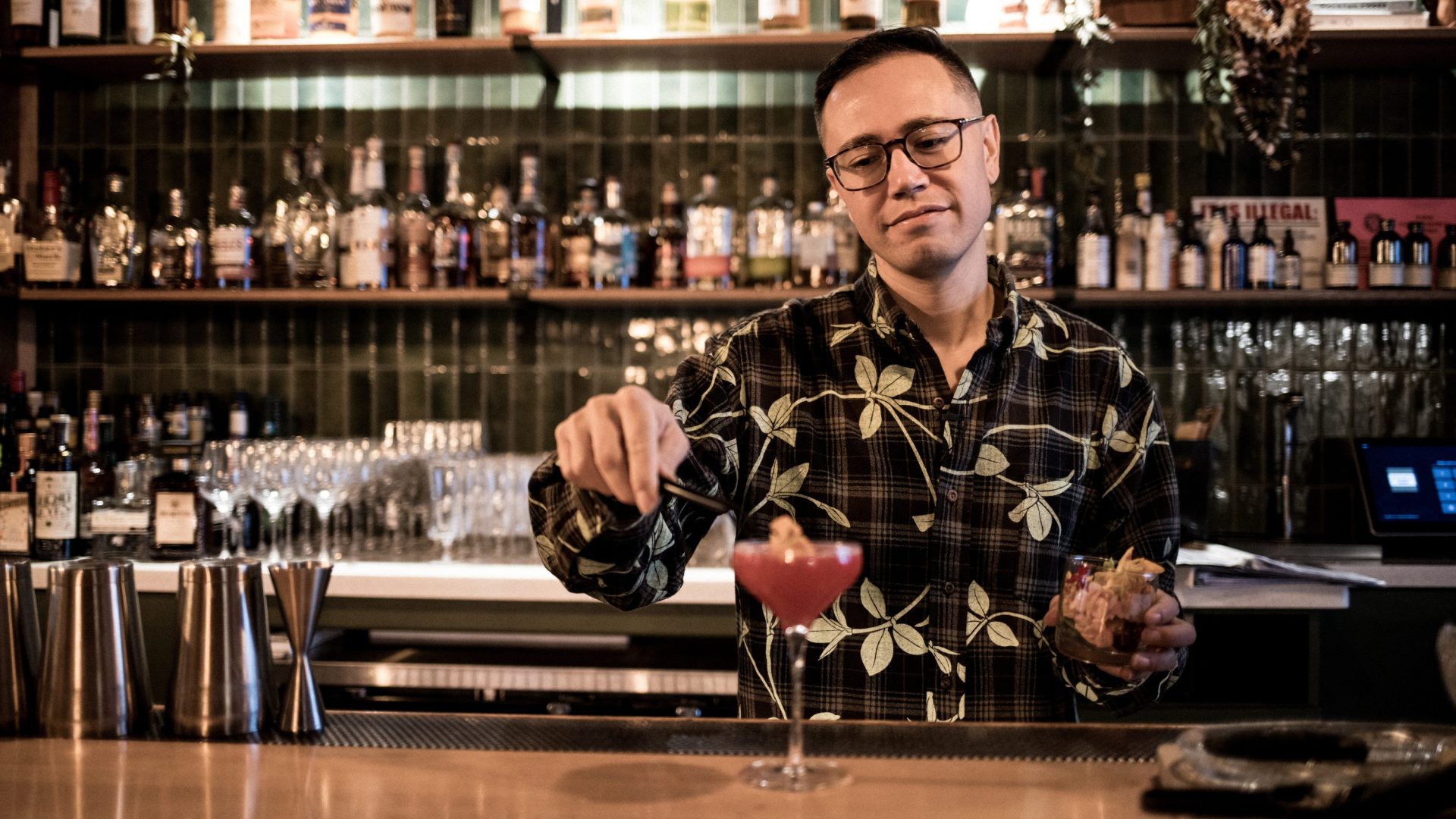 A man wearing floral shirt add edible flower to pink cocktail.