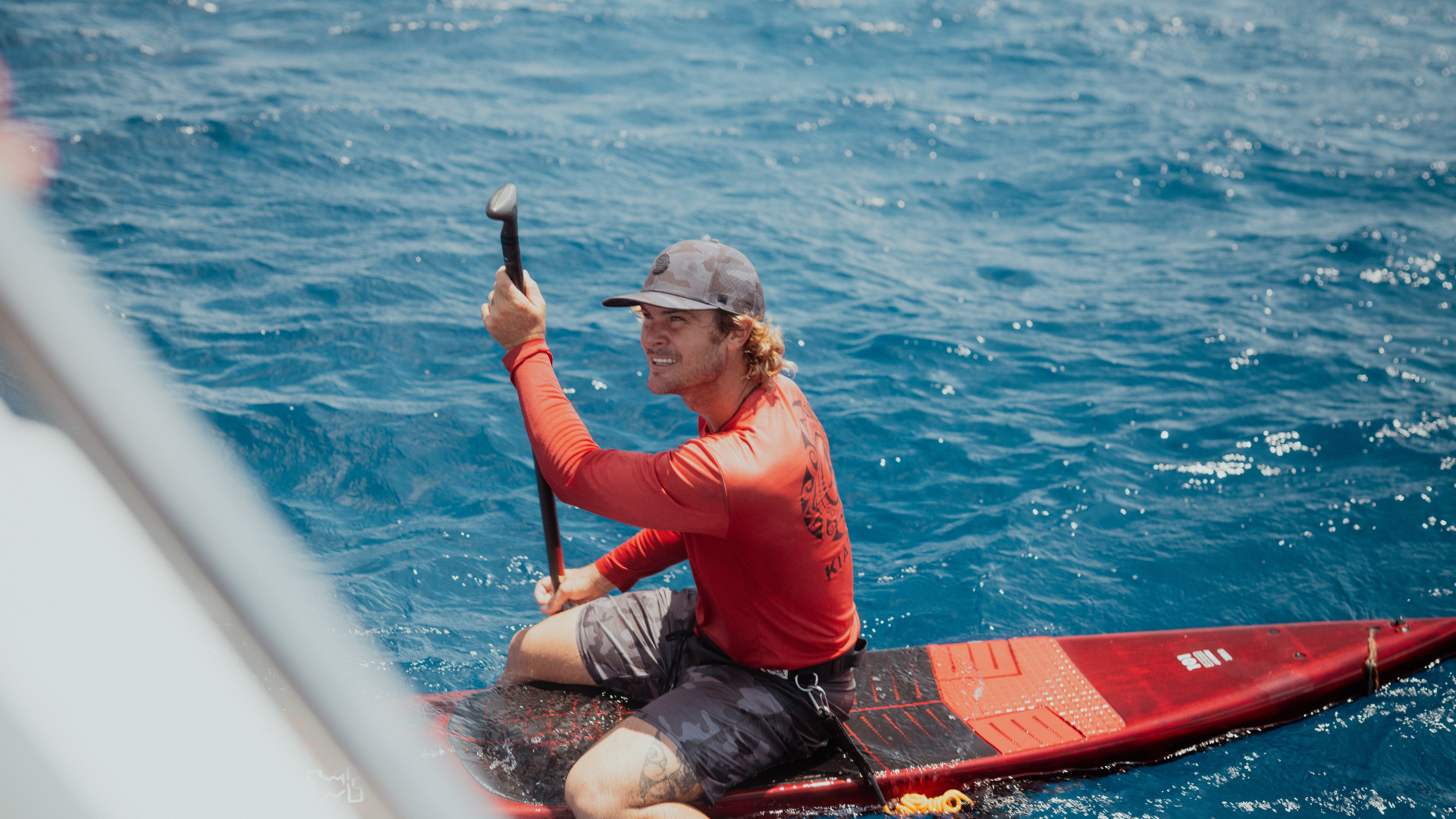 Man in red sits on hydrofoil board and uses paddle.