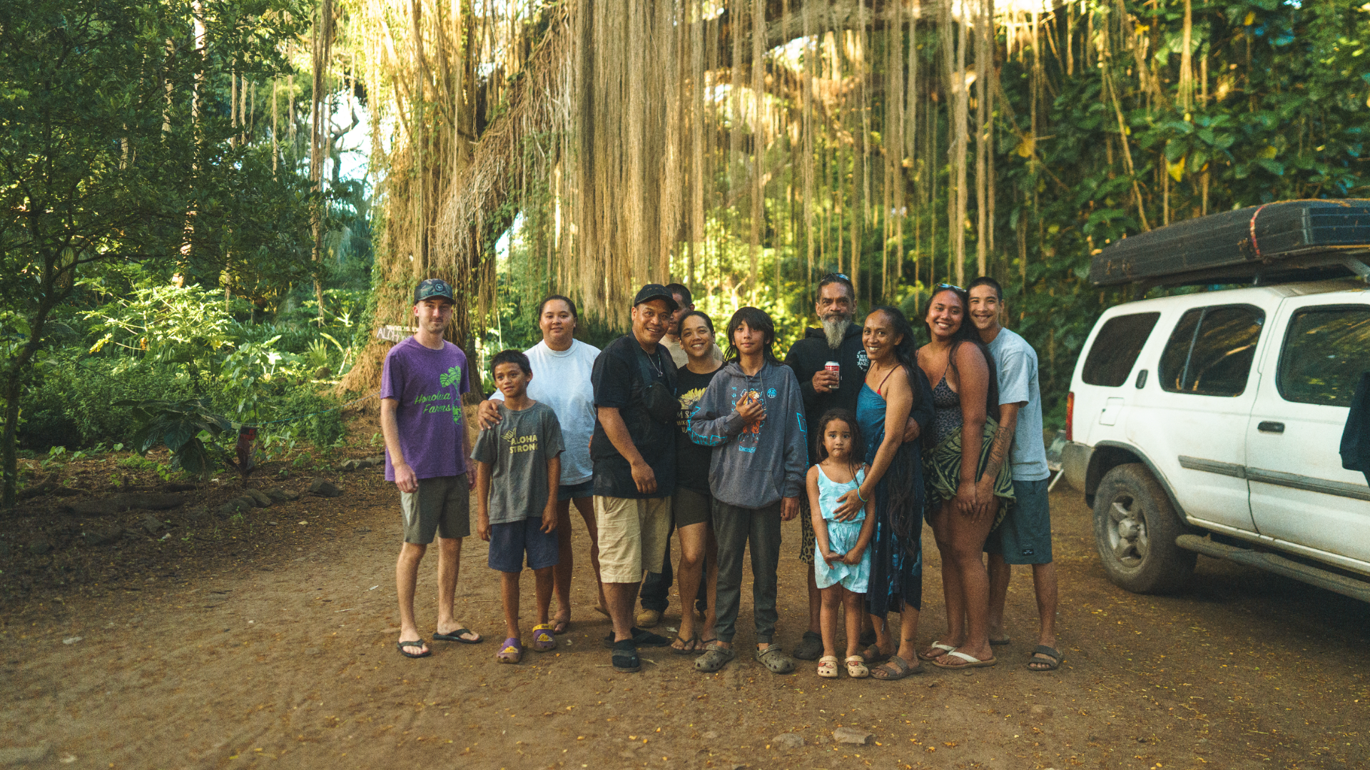 A family pose for a photo beside white car and beneath hanging vines.