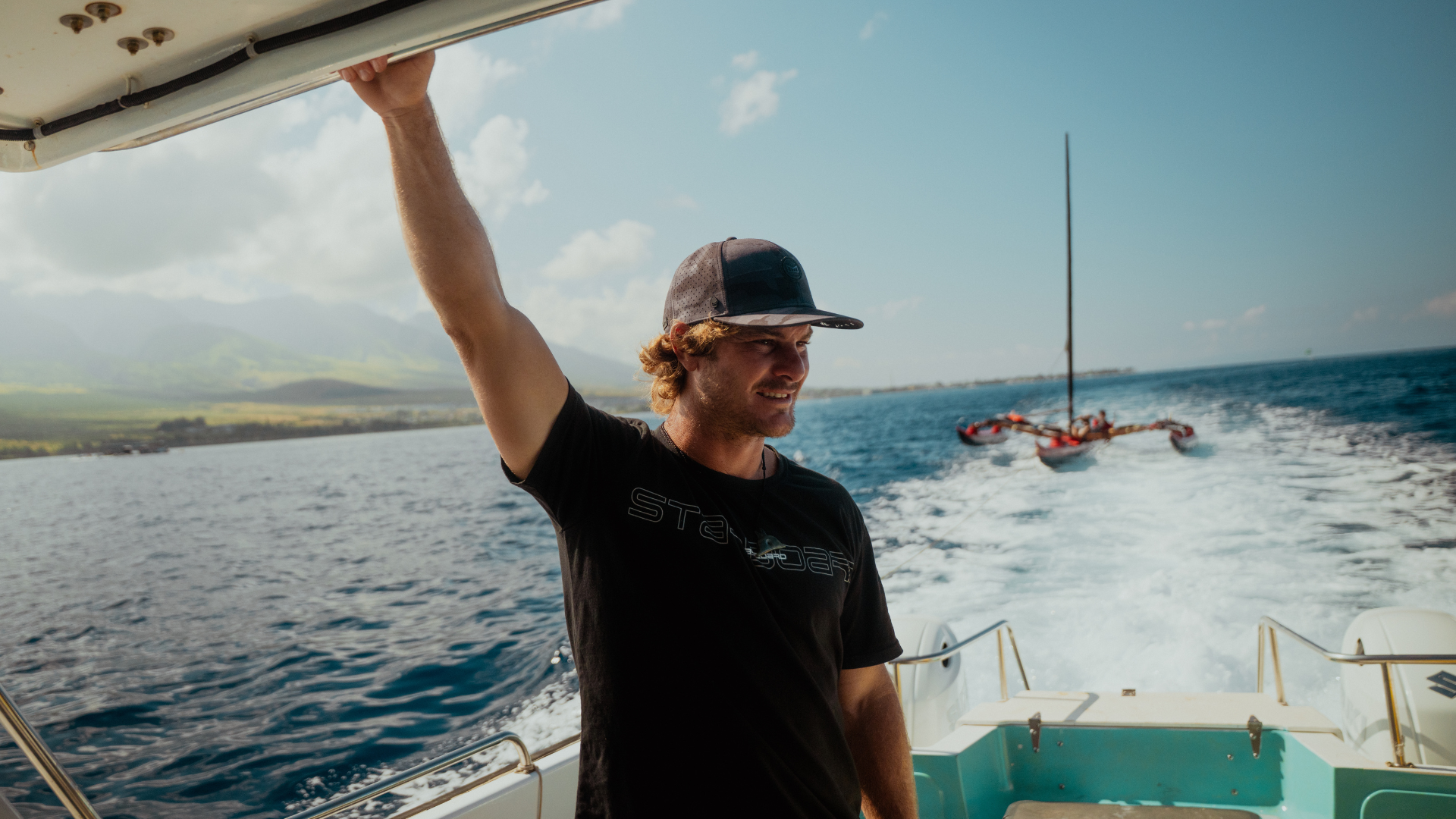 Man in black tshirt stands on boat with red sailing canoe behind him