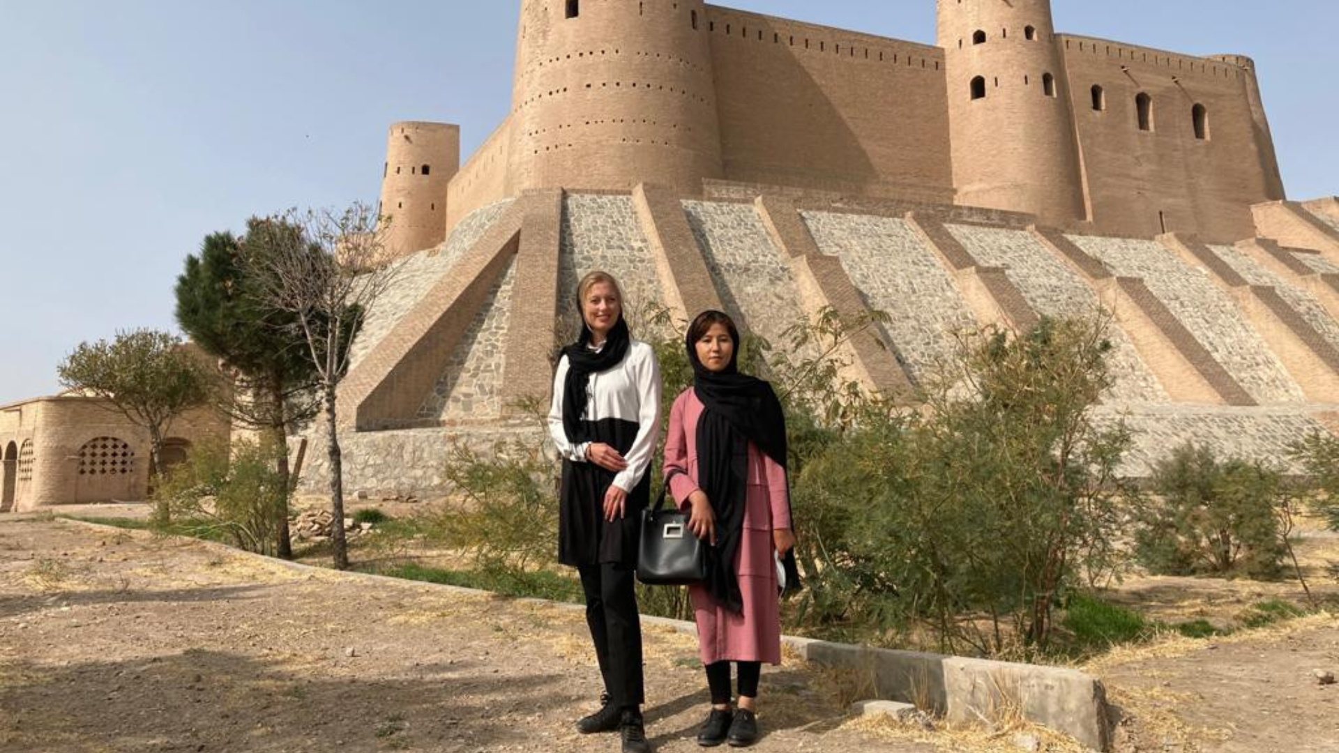 Two women wearing scarves stand infront of tan building.