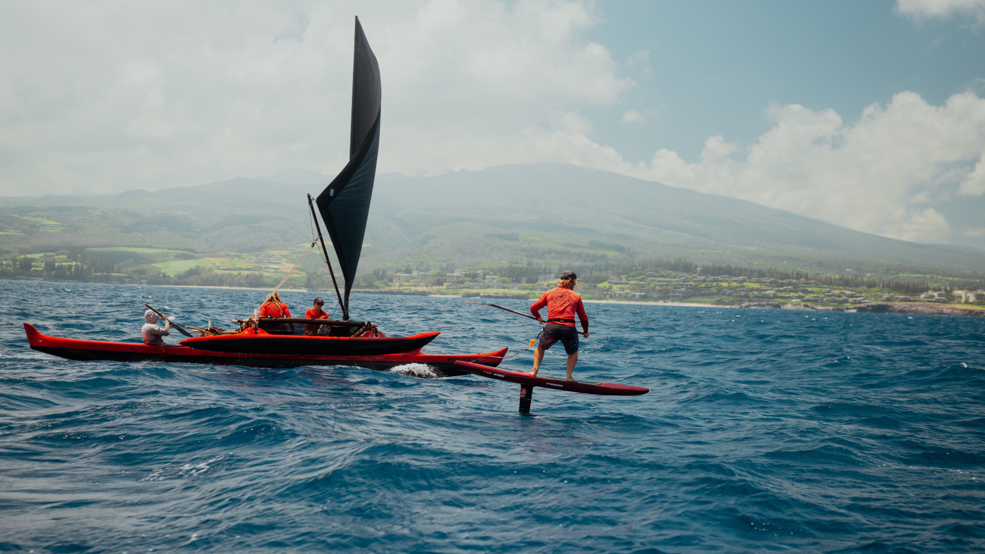 Man wearing red rides a red hydrofoil across water beside a red sailing canoe