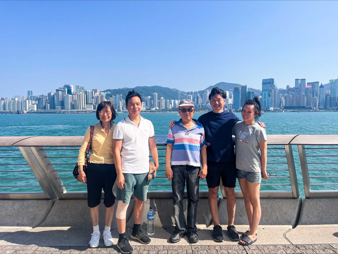 A family smiles for a photo in front of water with a city in the background.