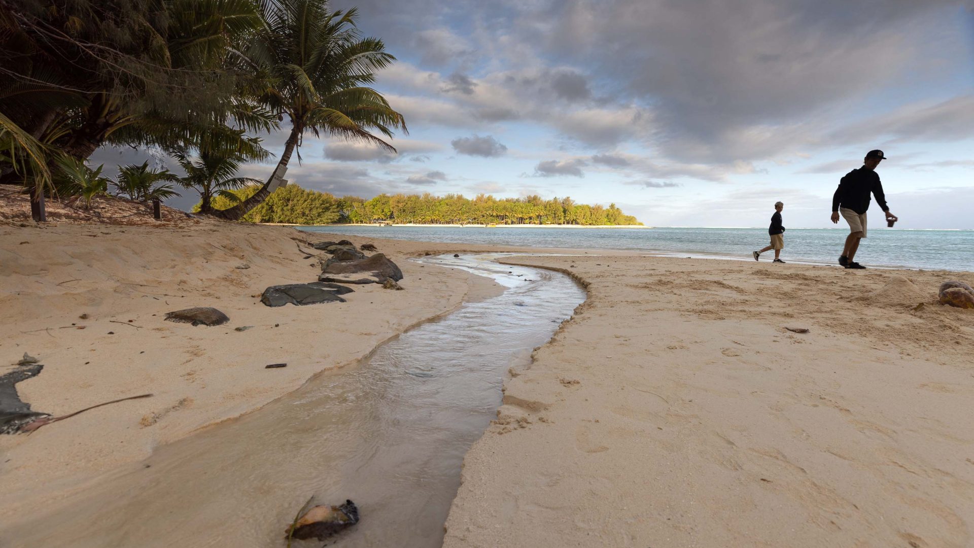 People walk along a beach with a stream running into it, and palm trees on the side. Clouds loom overhead.