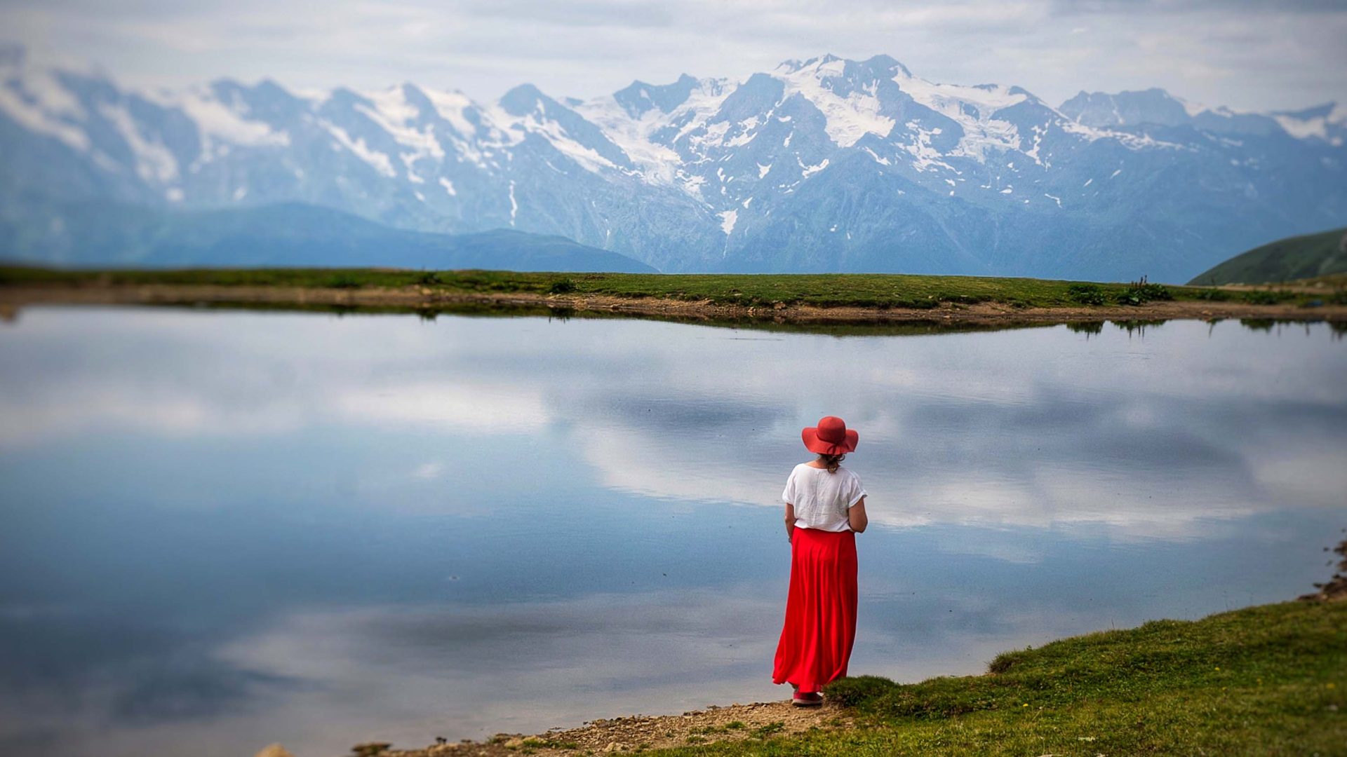 A woman in a red skrt and red hat looks out at snowy mountains behind a still lake.
