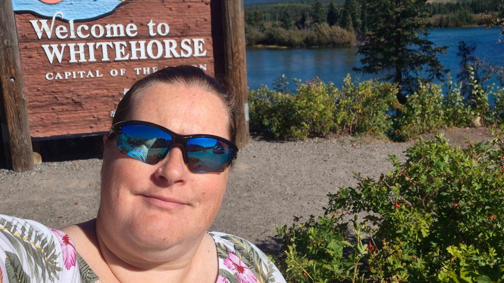 A woman in sunglasses takes a selfie in front of a sign that says 'Welcome to Whitehorse'.