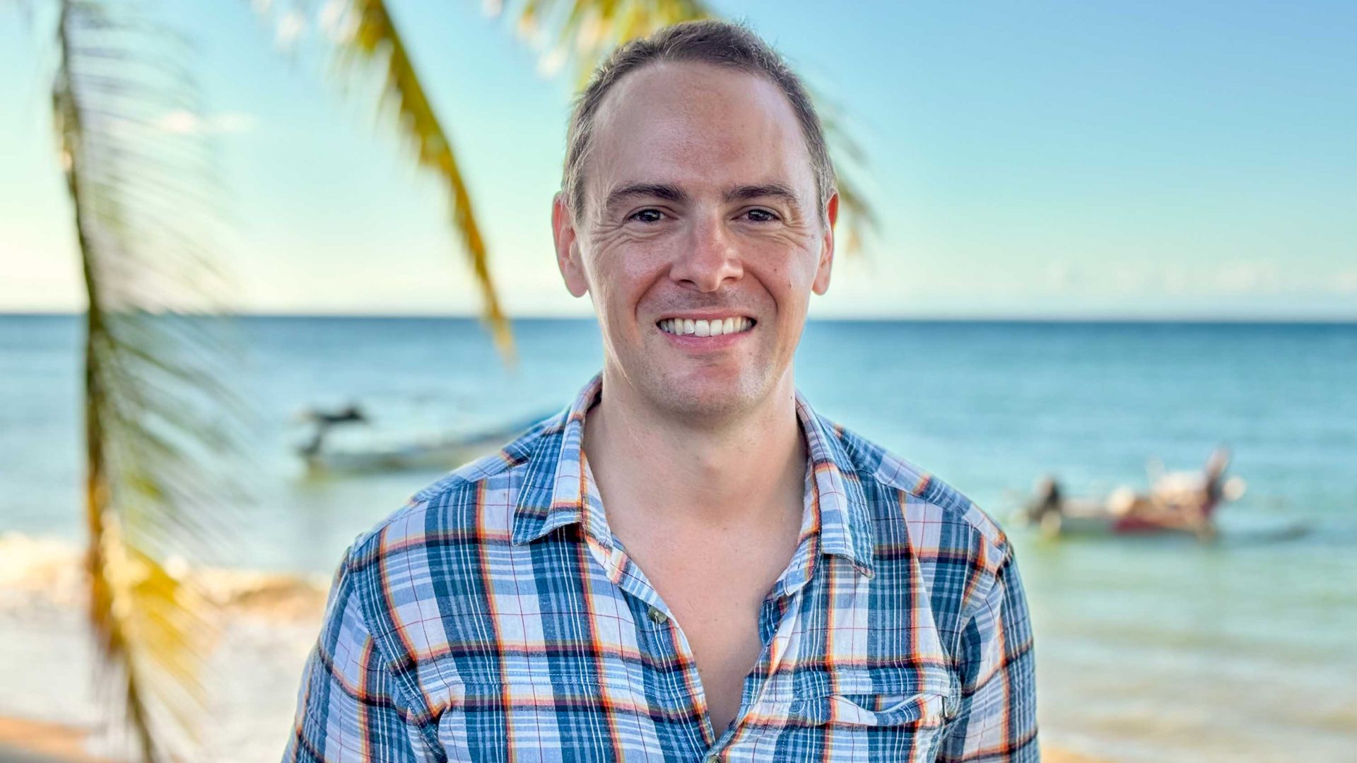A man smiles for a photo in front of a palm tree and sea.