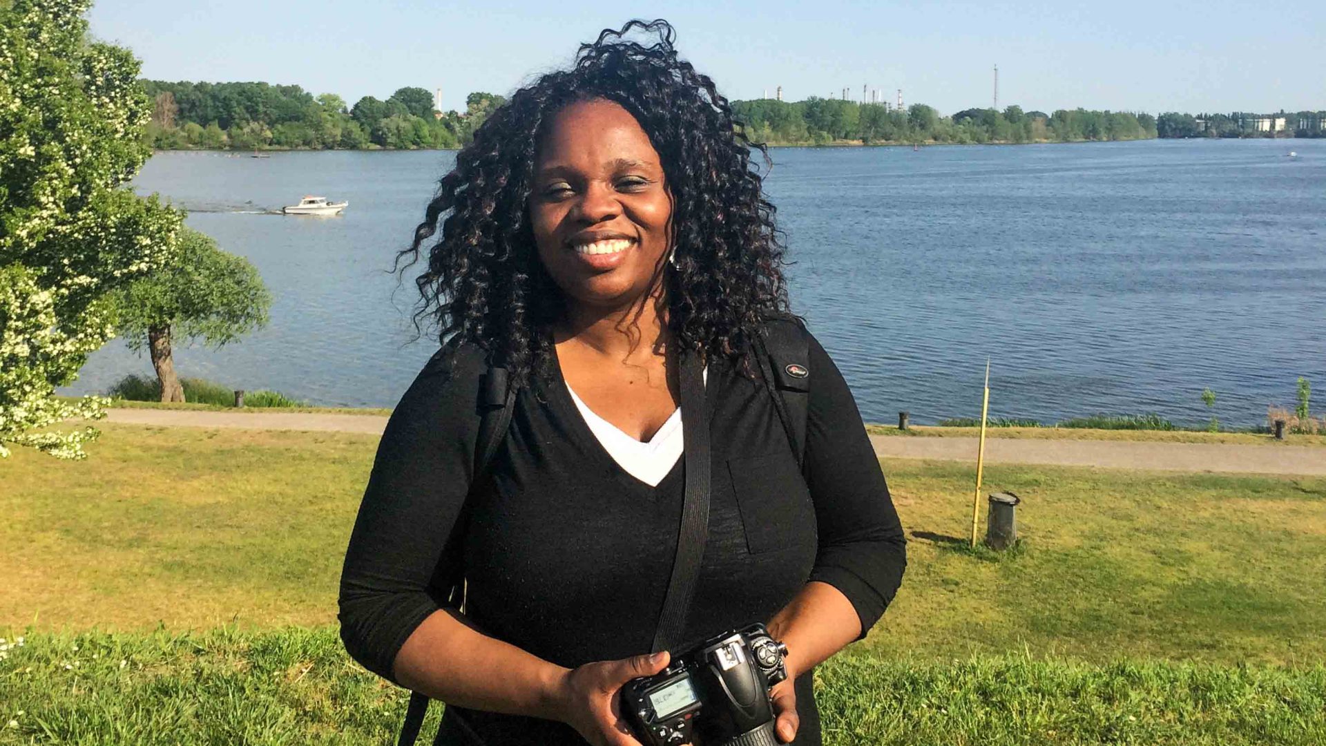 A woman with long curly hair smiles for a photo in the sun. She stands in front of water.