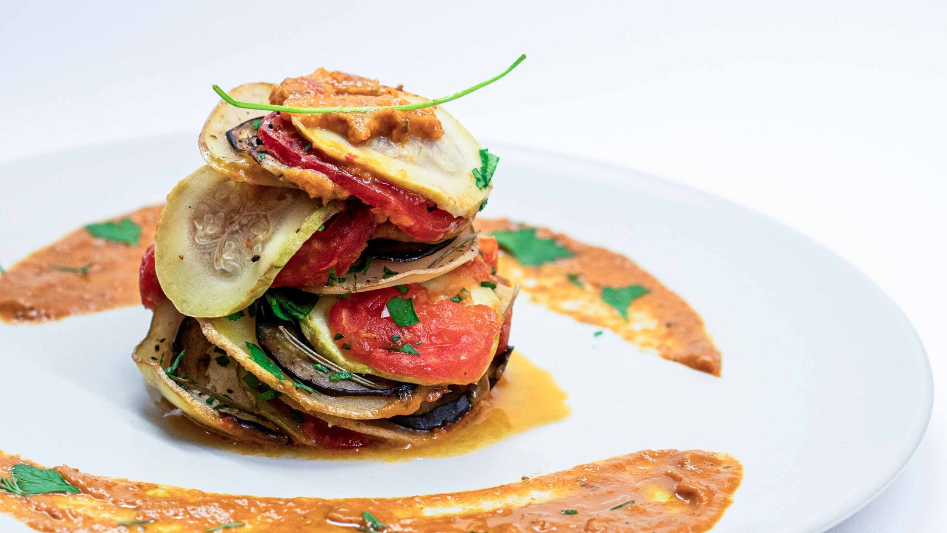 A white plate with a dish of layered vegetables with a sauce around the edge of the plate.