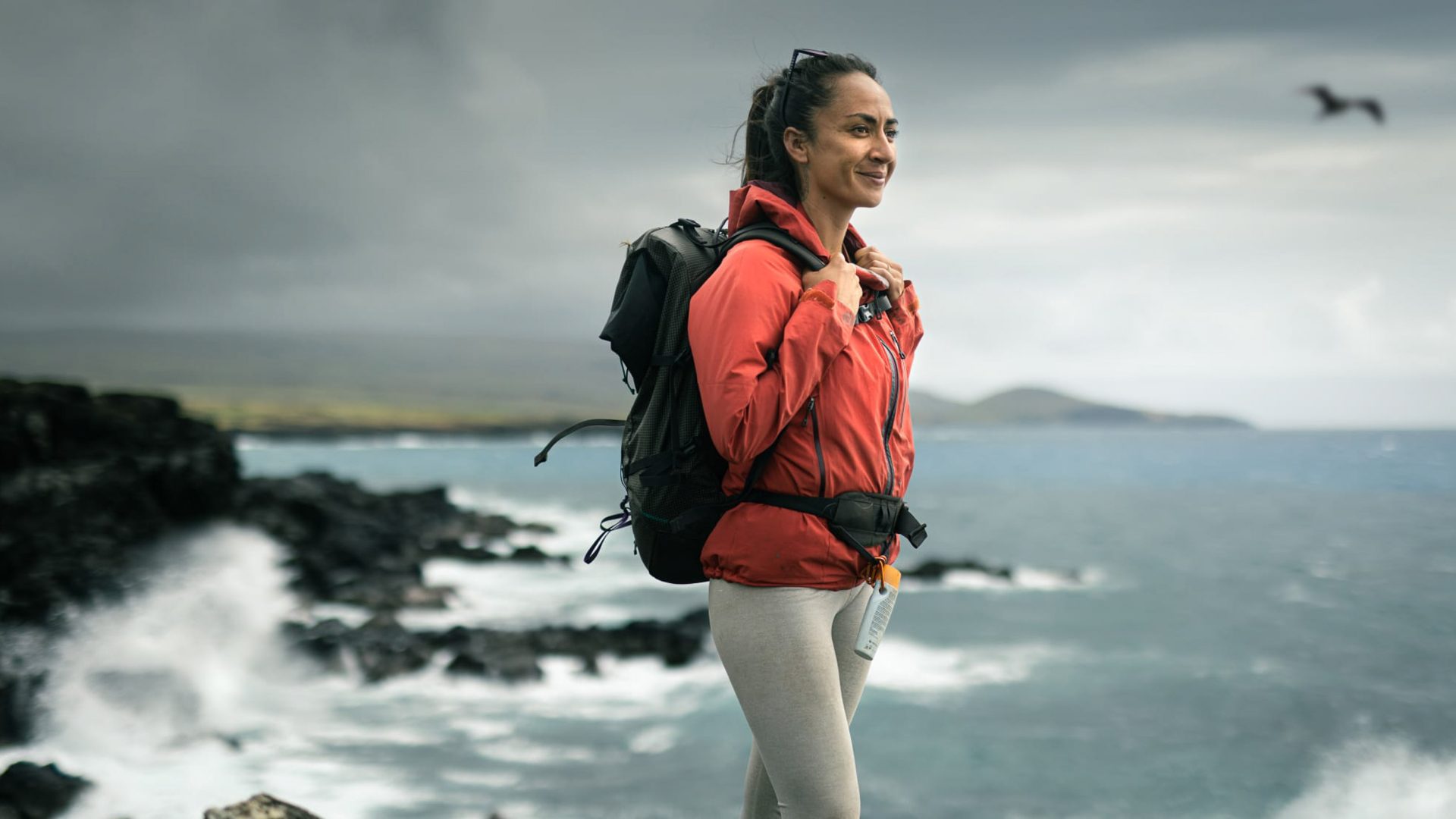 A woman in an orange jacket and backpack stands near the coast.