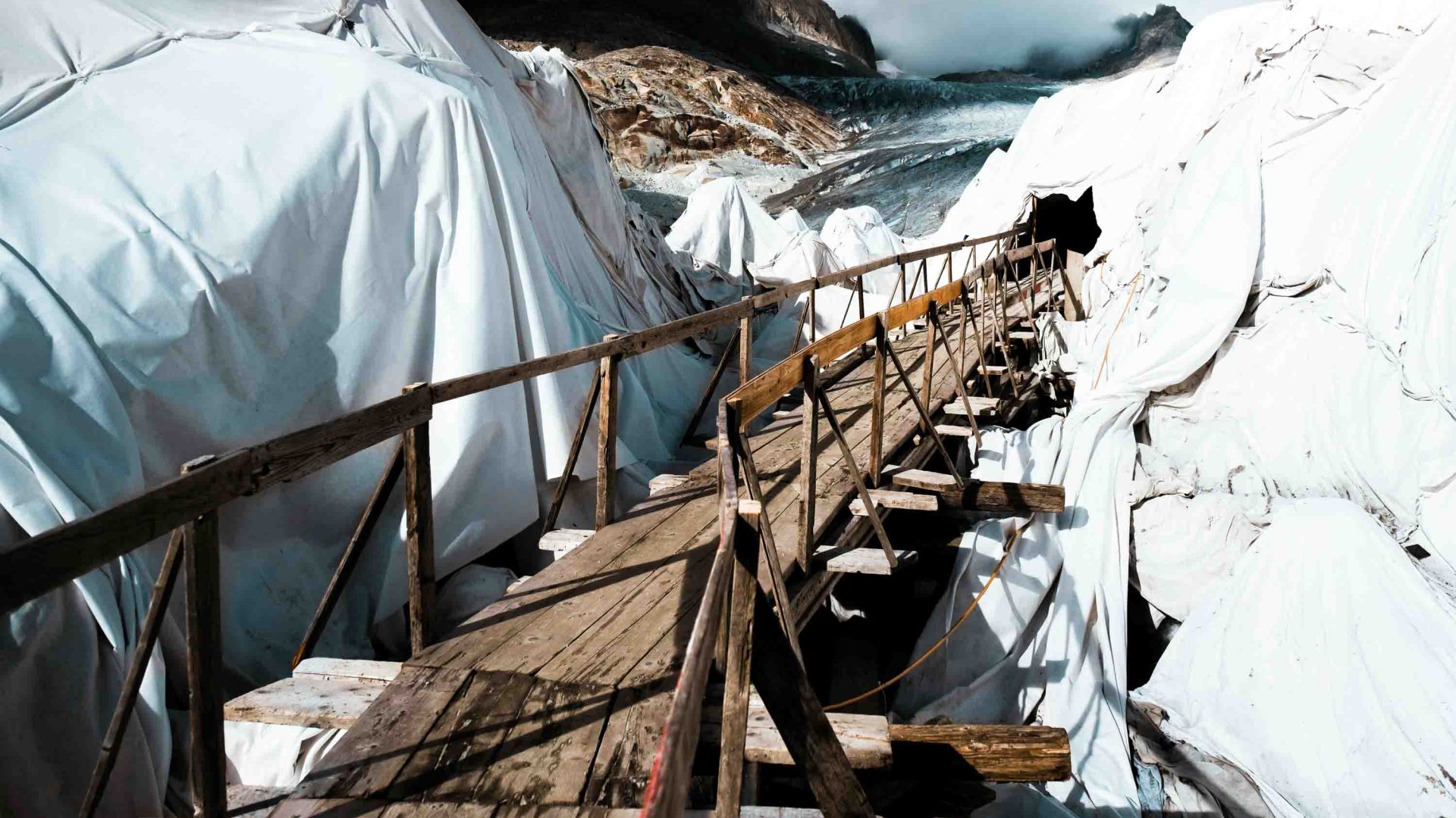 A rickety wooden bridge passes through glaciers covered in white sheets.