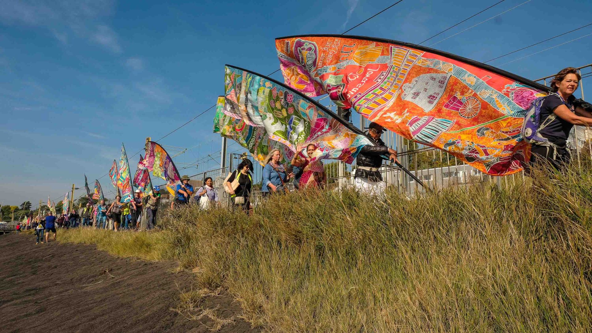 People holding long silk flags walk past long grass on a trail.