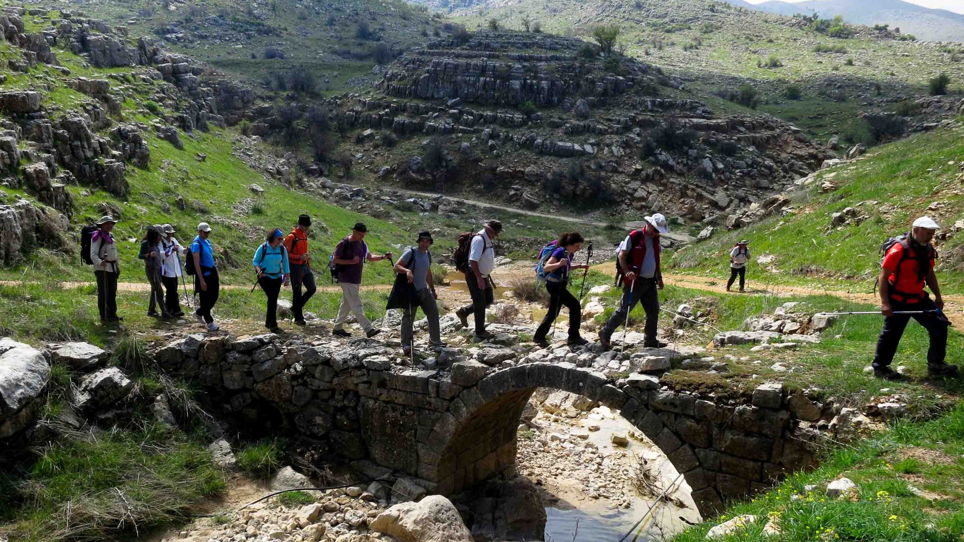People hike in a row over a stone arched bridge.