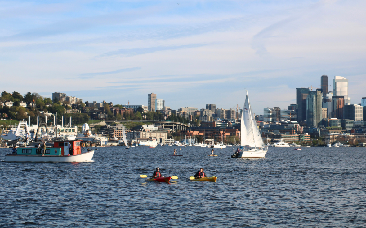 Two kayakers cross water infront of city and sailboats