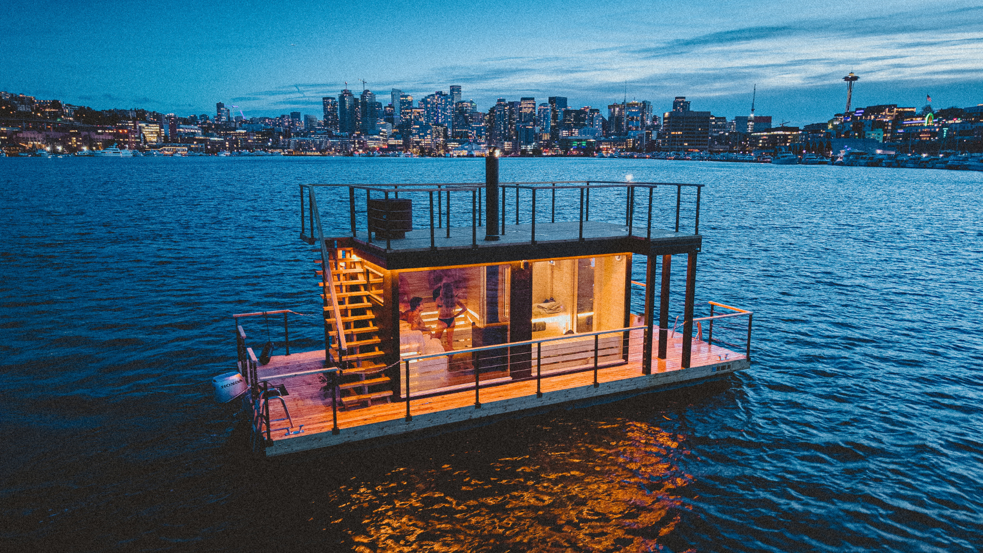 Sauna lit up from inside, floating on dark water in front of Seattle skyline
