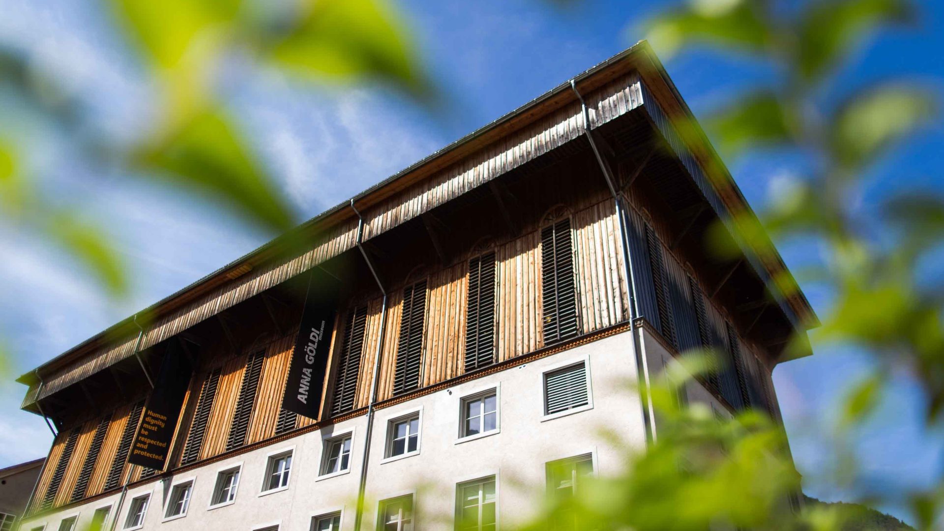 A building with the words "Anna Göldi" on it is seen through the leaves of a tree.