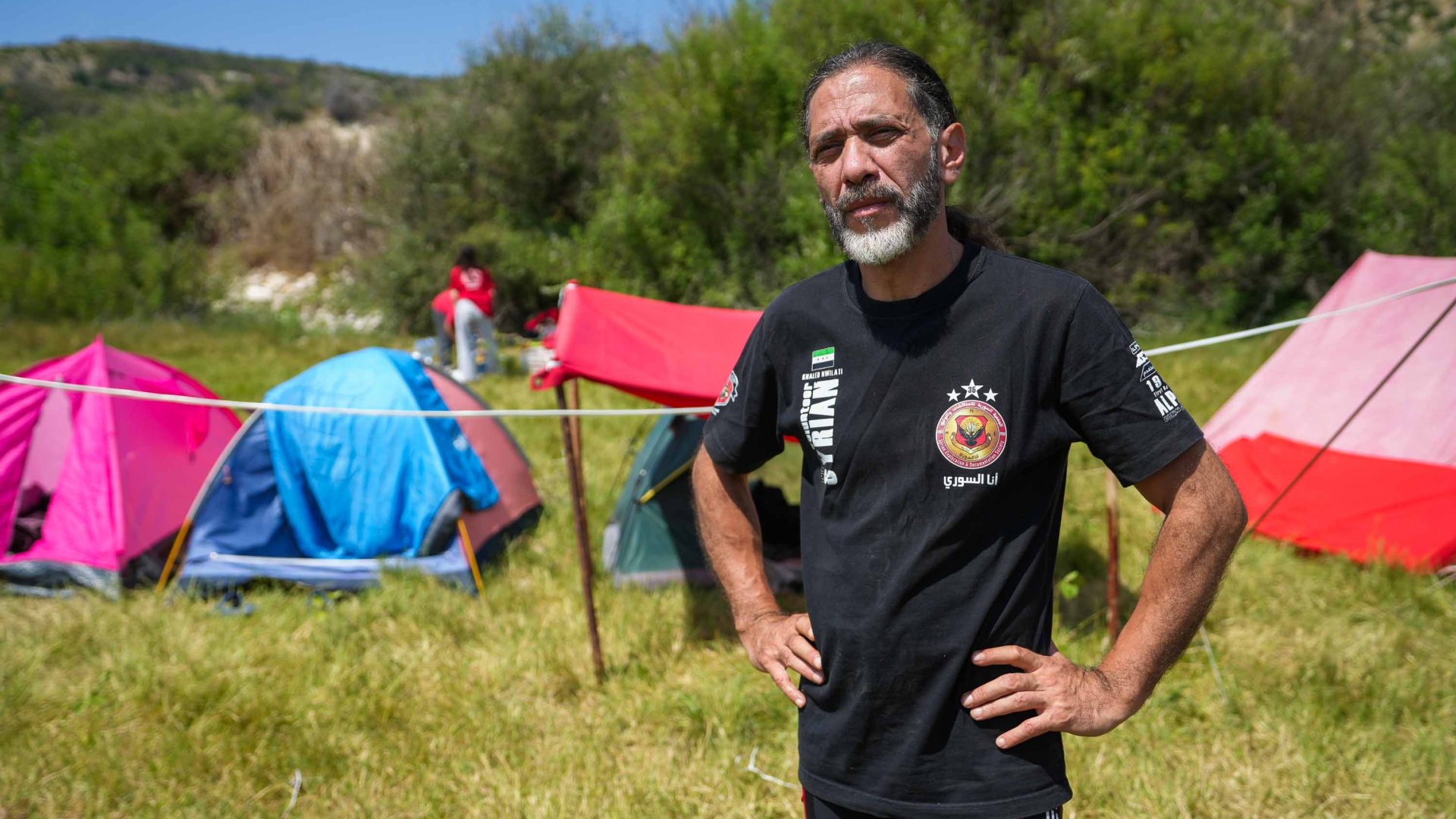 A man with hands on hips stands in front of some tents.