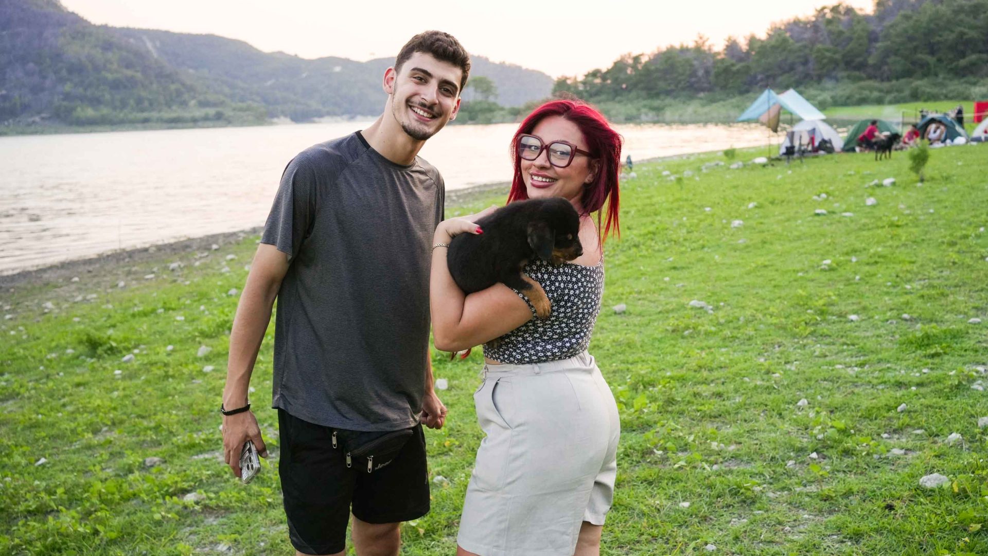 A young man and woman pose for a photo at a campsite. The girl is holding a black puppy.