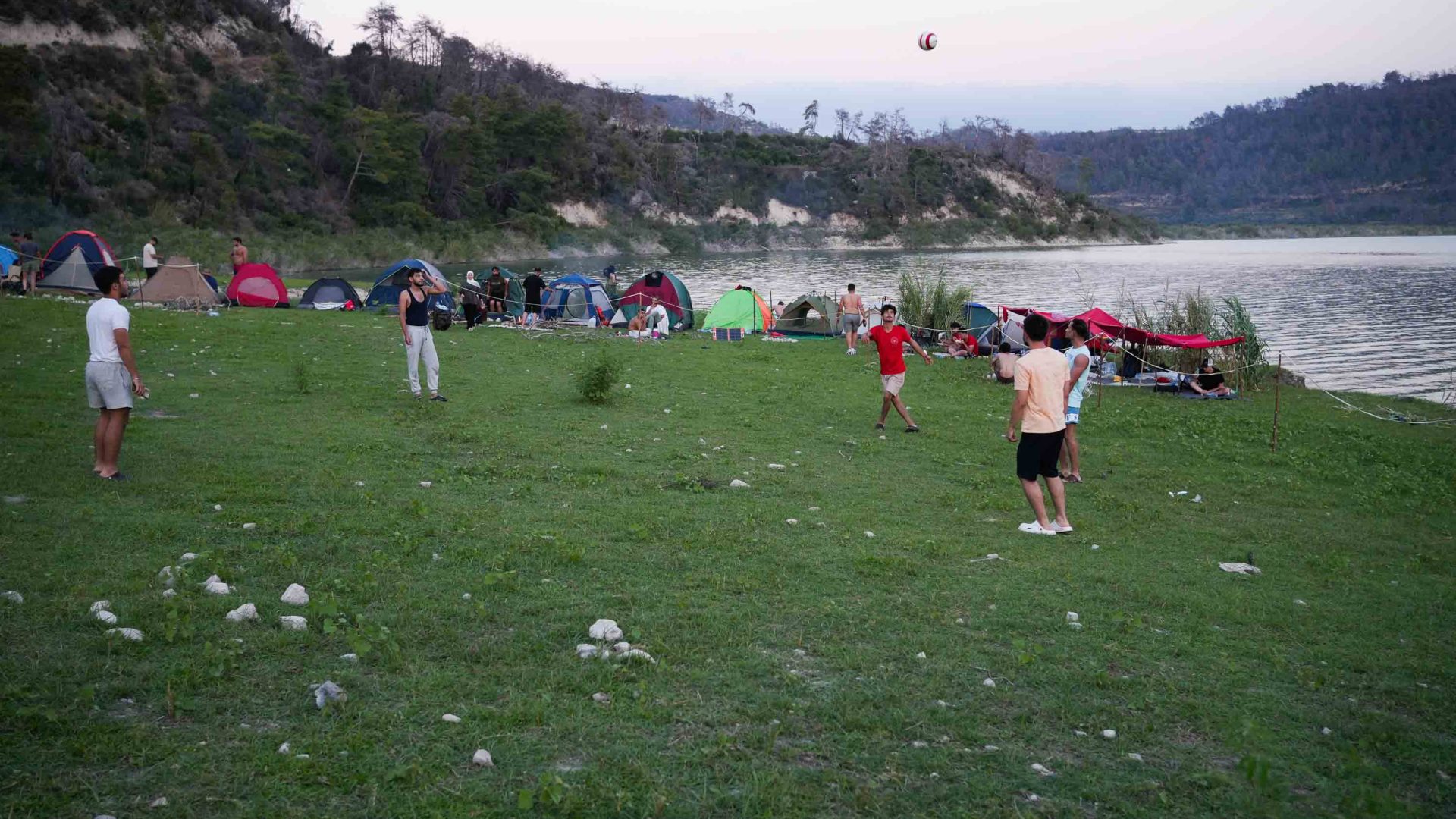 People play soccer by a lake, with tents in the background.