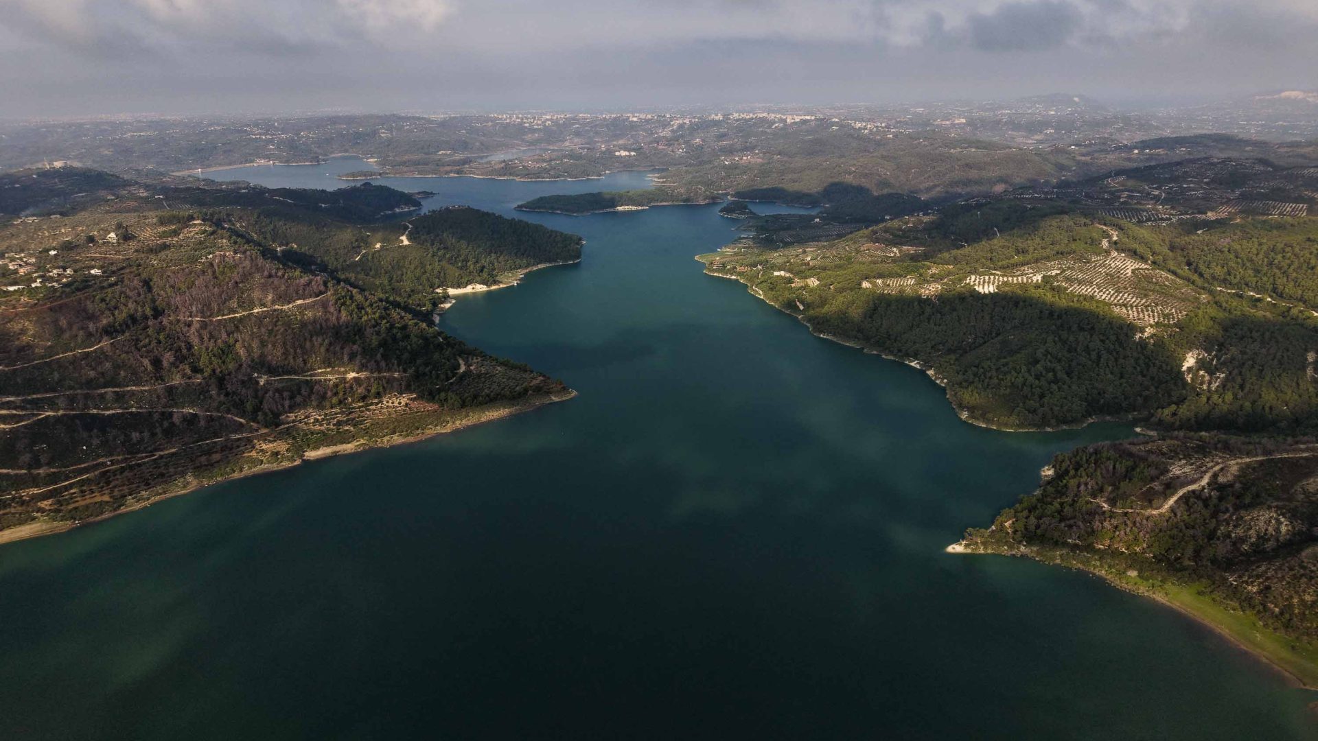 A drone photo of pine covered land bordering a deep blue lake.