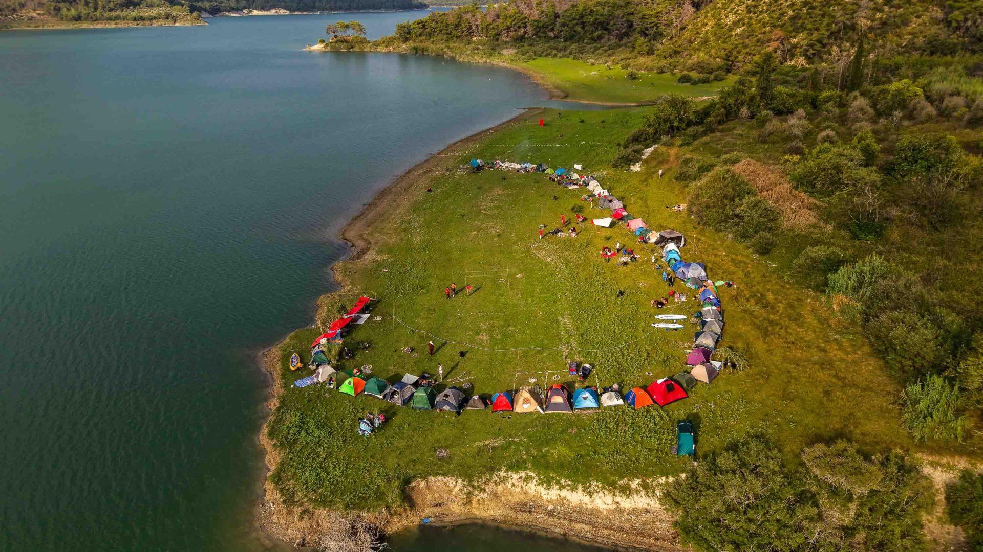 Aerial views show the campsite and surrounding landscape, with tents set up along the shore of the lake and mountains stretching beyond.