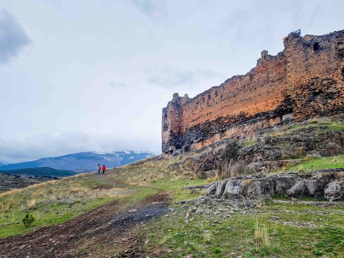 An old stone castle stands on the top of a mountain where two hikers can be seen walking.